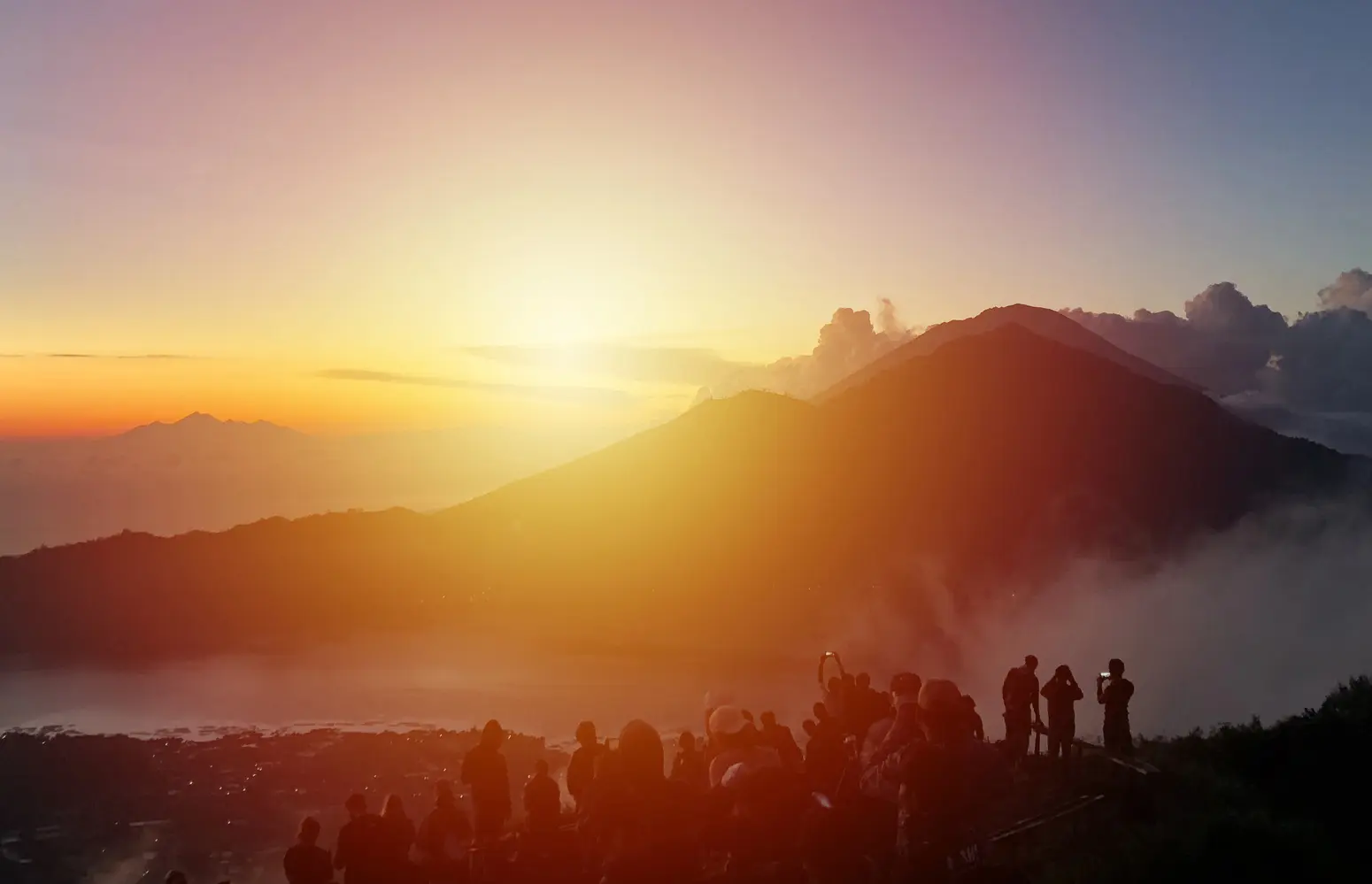 A group of people standing on Mount Batur watching the yellow and orange sunrise over the mountains, one of the top things to do in Bali.