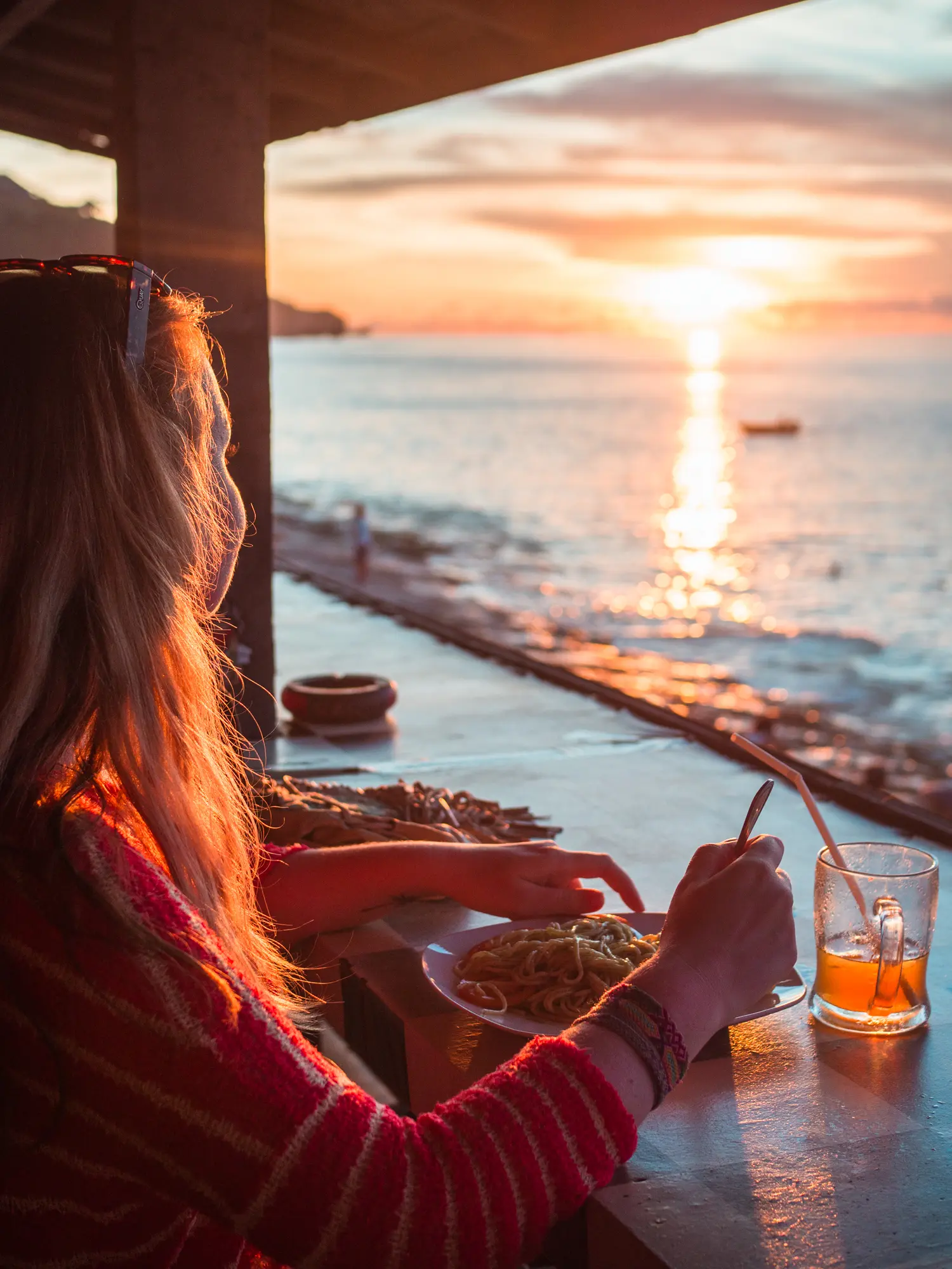 Woman wearing a red sweater sitting at a table eating noodles looking out at the sunset at Bingin Beach in Bali vs. Lombok.