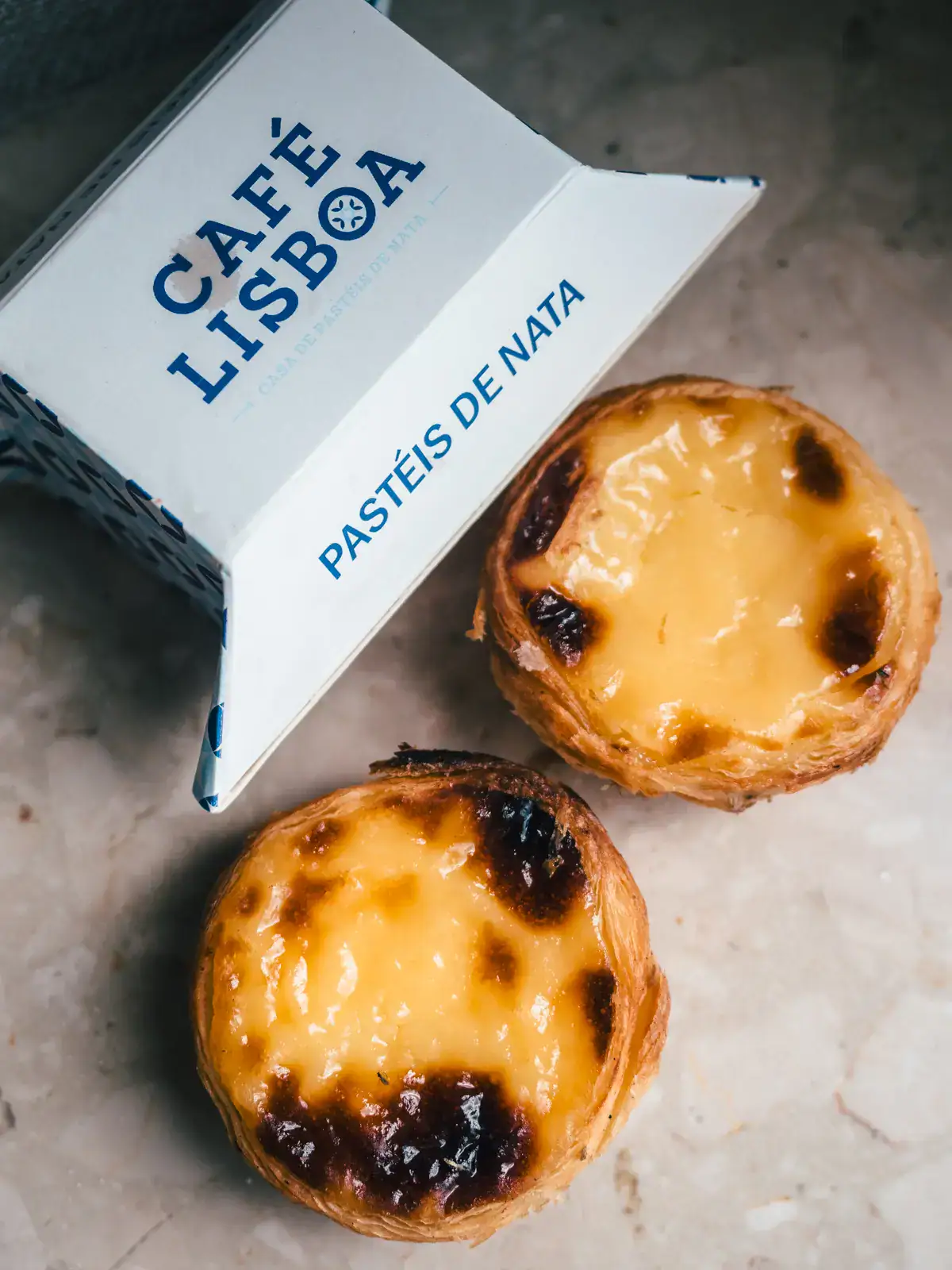 Two round, baked custard tarts - Pasteis de Nata next to a white and blue Café Lisboa takeaway container, one of the best pastries in Krakow.