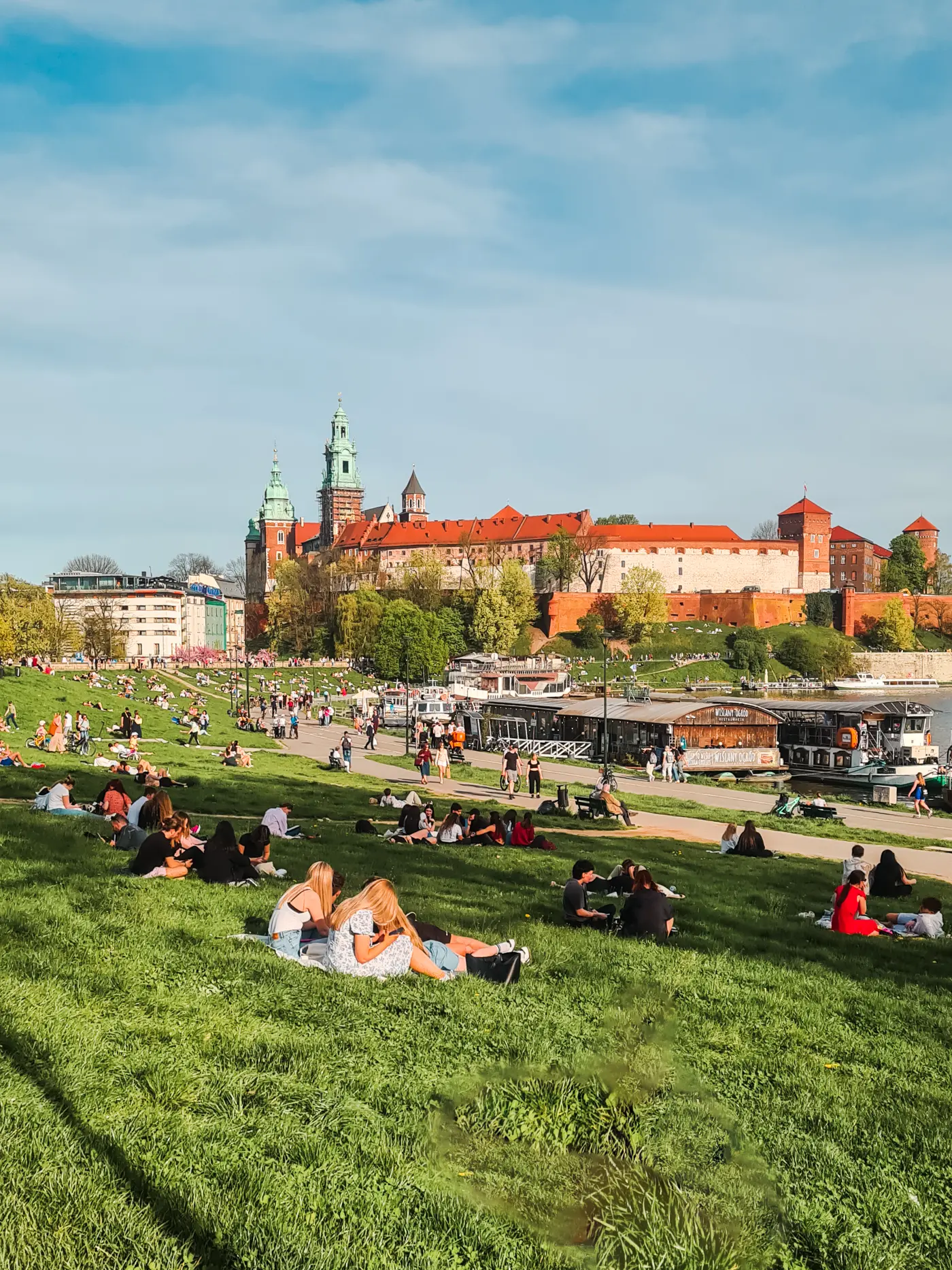 People enjoying the sun on the green lawn along the river with Wawel Castle in the background, on a 2-day weekend in Krakow.