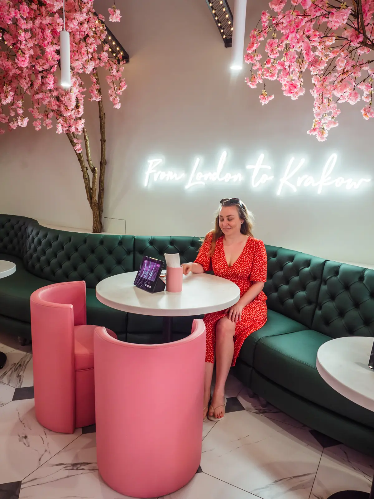 Woman in a red dress sitting on a green leather sofa by a white table and two pink chairs with pink cherry blossoms in the ceiling at EL&N London, on a weekend in Krakow.