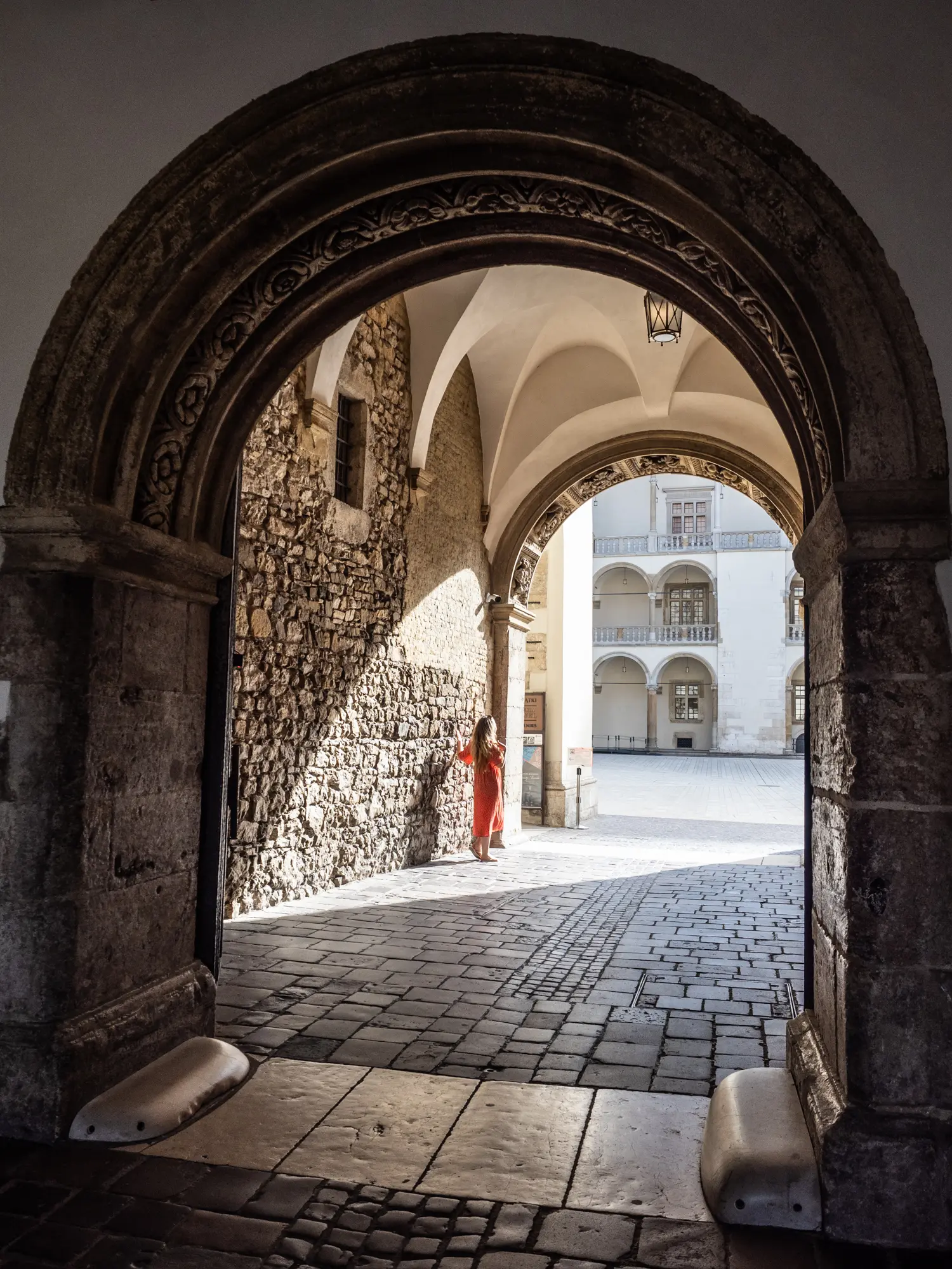 Woman wearing a red dress standing in an arched hallway looking out over the sunny courtyard at Wawel Castle in Krakow.