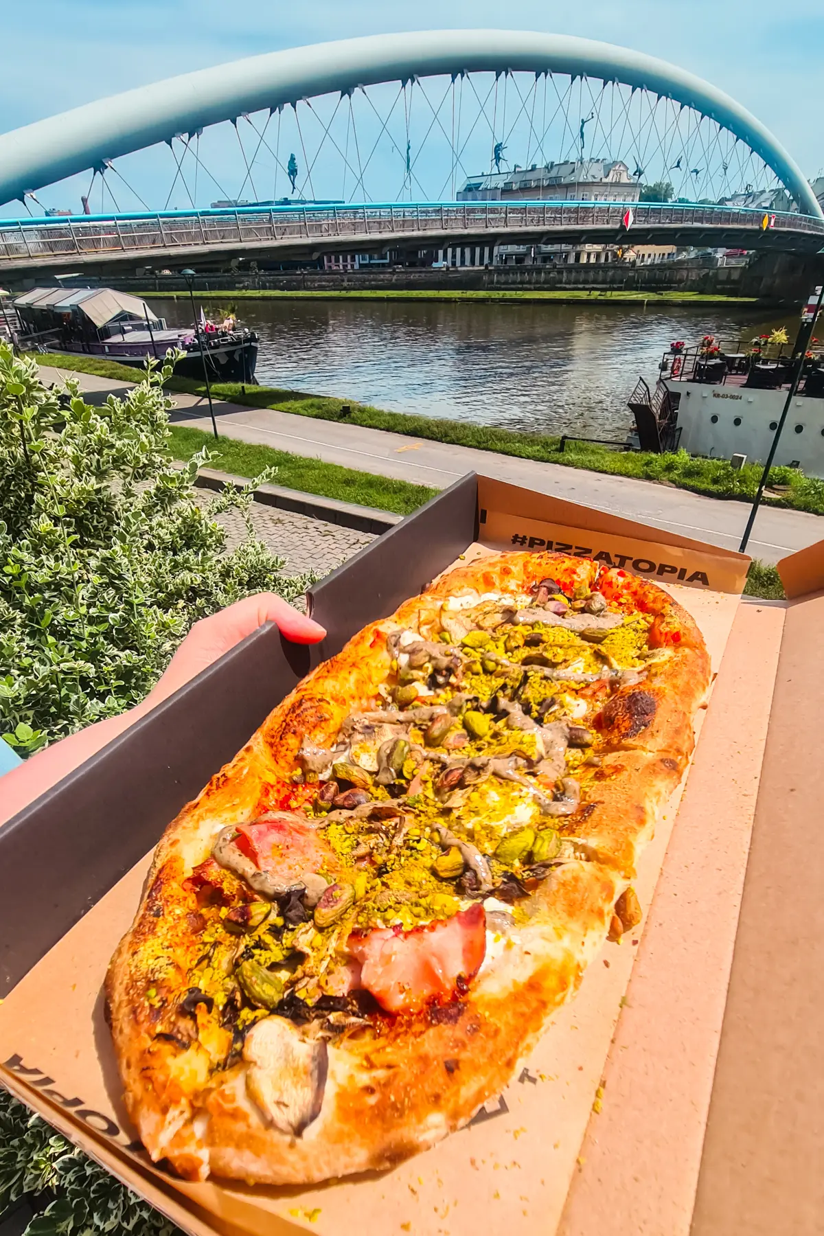 Long pizza topped with pistachio nuts in a cardboard box in front of a bridge by the river, during a weekend in Krakow.