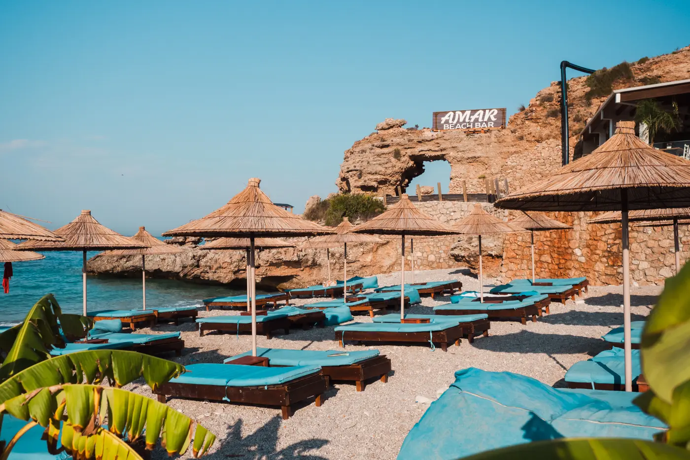 Blue sunbeds, straw parasols on a pebble beach with a stone arch in the background in Dhermi Albania, one of the best summer destinations in Europe.