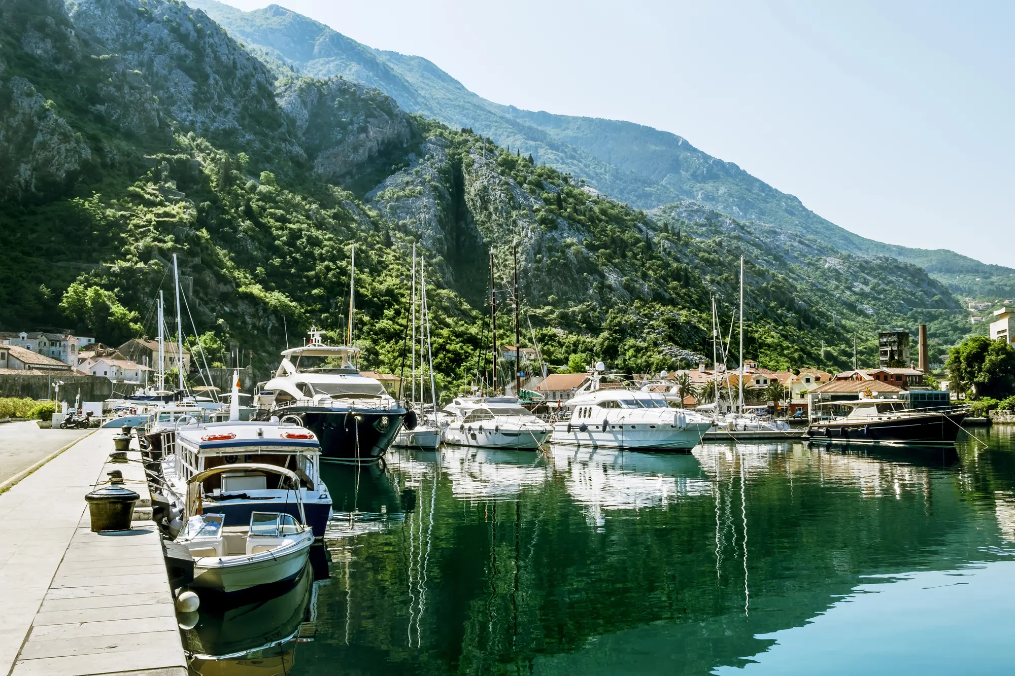 White boats and a black yacht in the calm harbor in Kotor with mountains in the background and green water, one of the best summer destinations in Europe.