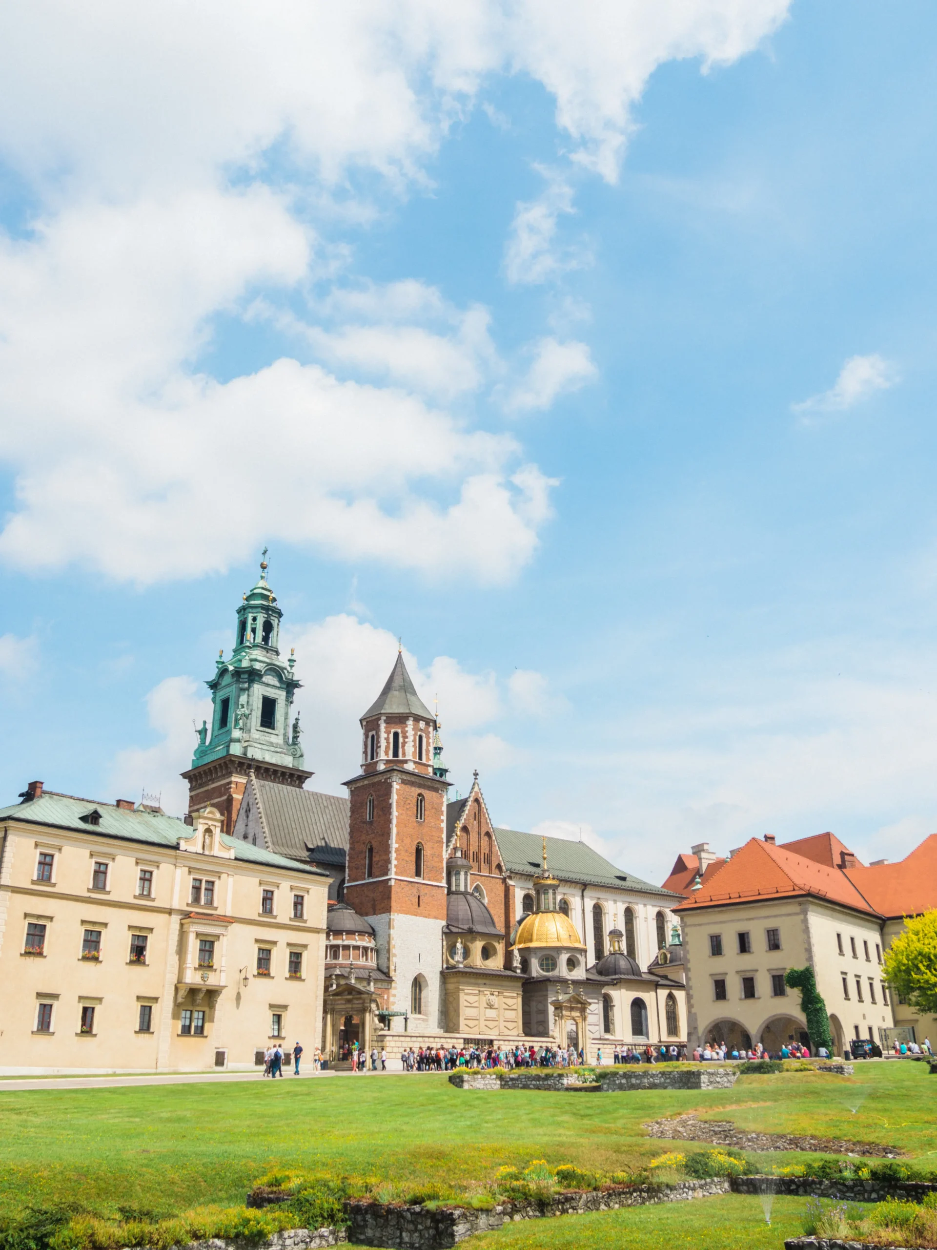 Wawel Castle and Cathedral with many different architectural styles and towers, green lawn in front and people walking.