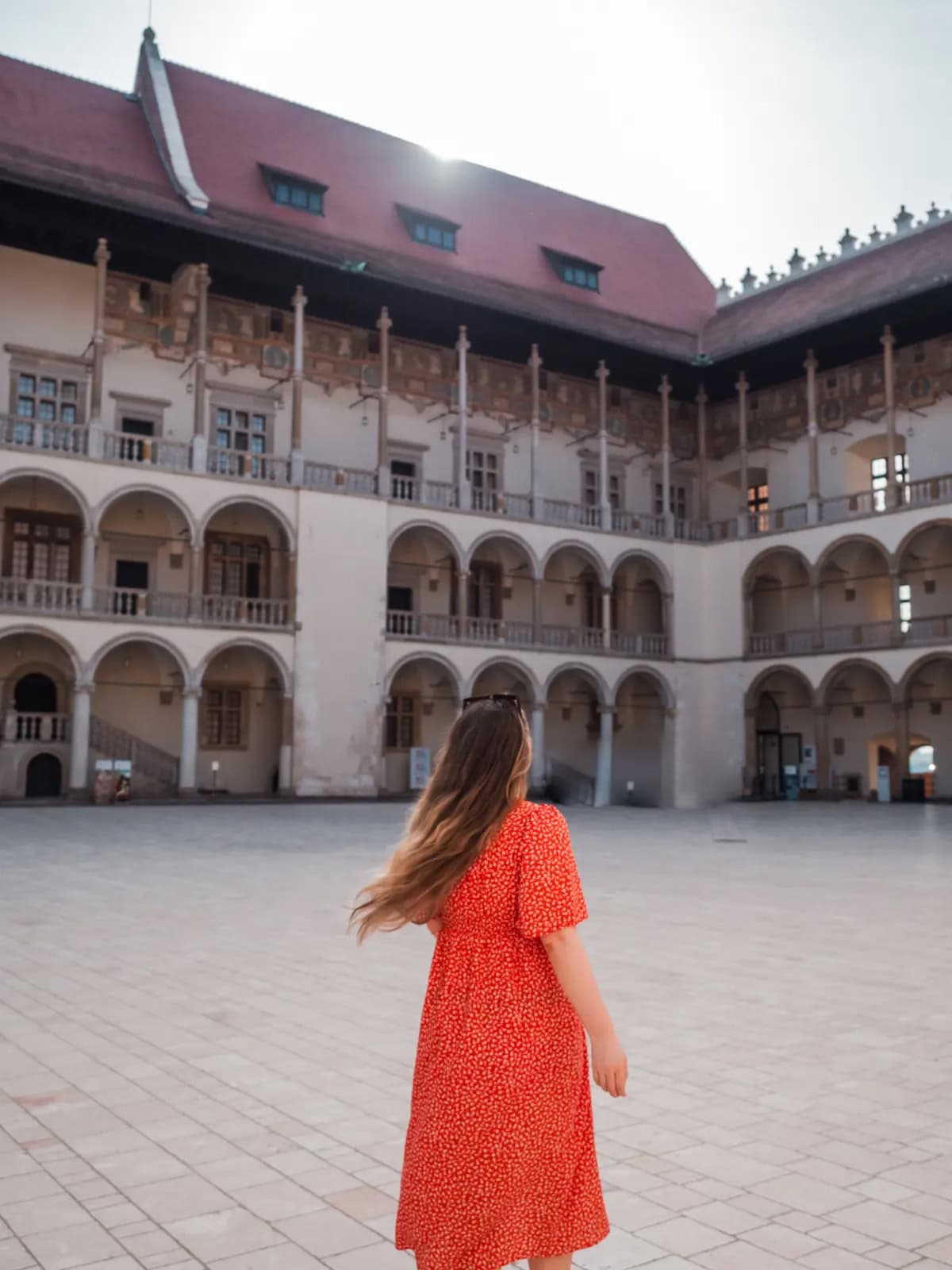 Woman with a red dress and long hair walking in the large courtyard of Wawel Castle with several levels with archways in Krakow.