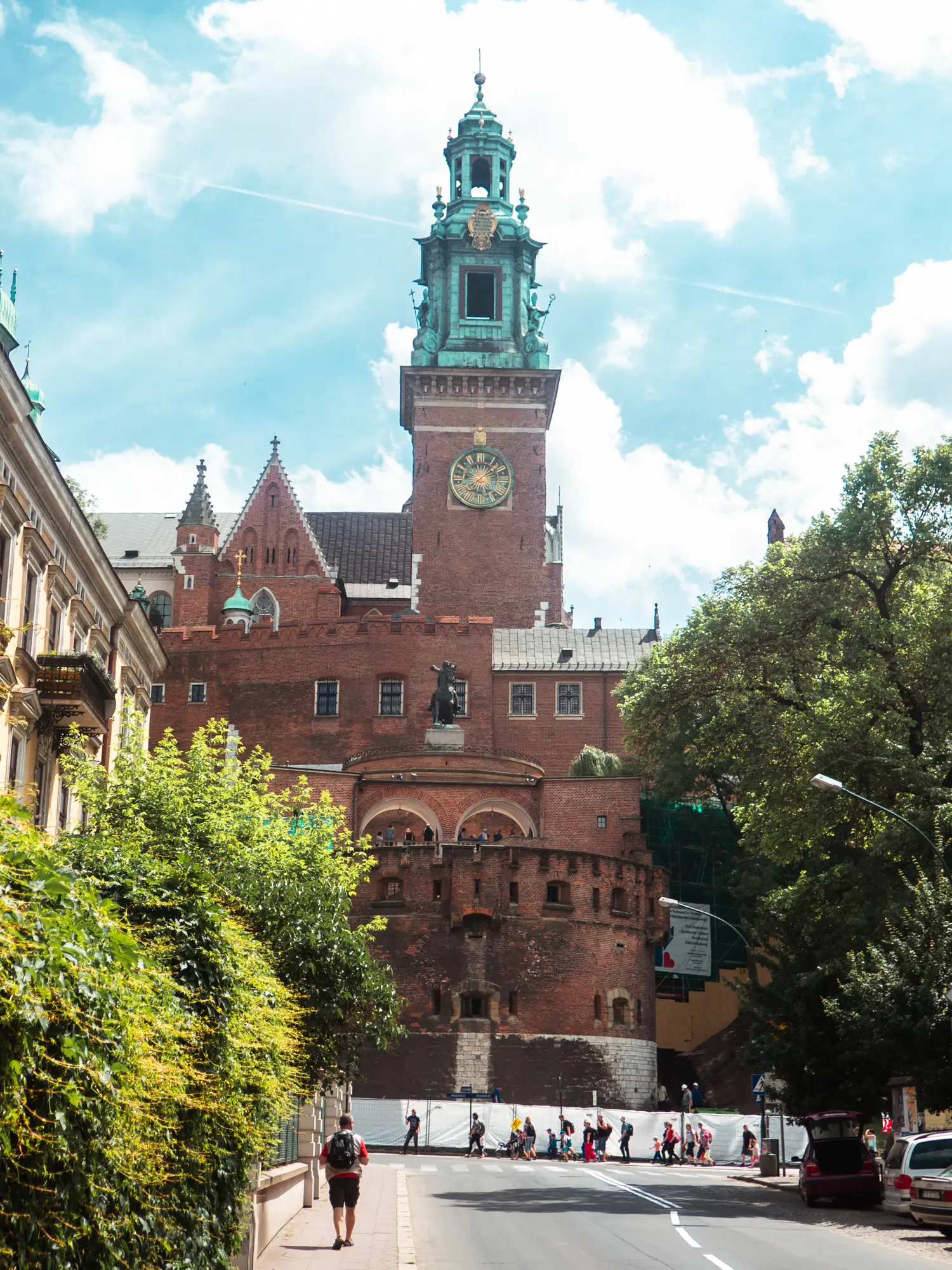 Walking up the street towards the tall clock tower of Wawel Castle made from red brick, with green trees of both sides.