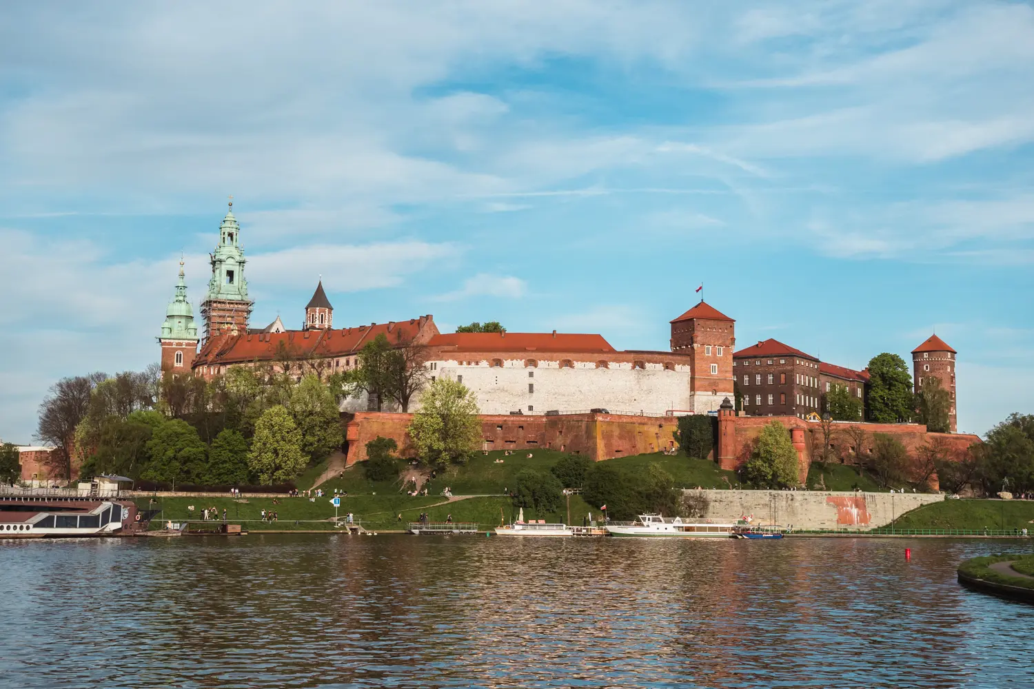 Red brick Wawel Castle surrounded by greenery seen from a river cruise on Vistula River.