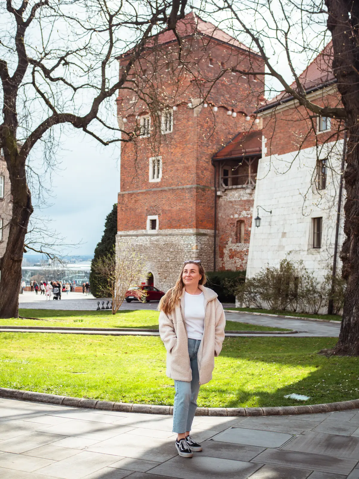 Woman wearing jeans and a white coat on the grounds of Wawel Castle in Krakow with a green lawn and a brick tower in the background.