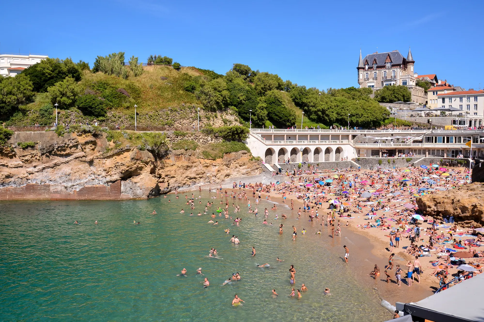 Beach full of people with green water and old buildings and greenery in the background in Biarritz, one of the best summer destinations in Europe.