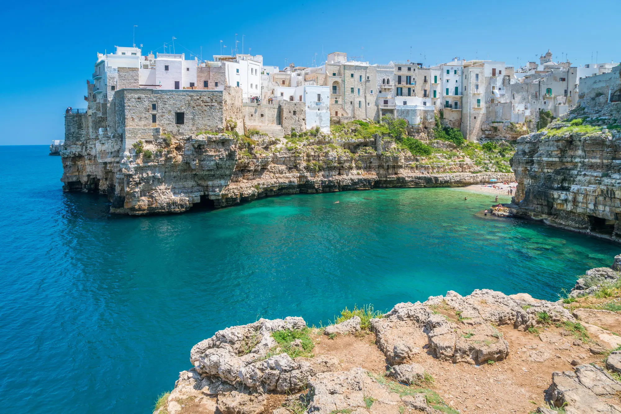 Old buildings on an outcrop overlooking a beach and turquoise ocean in Polignano a Mare in Puglia, one of the best summer destinations in Europe.