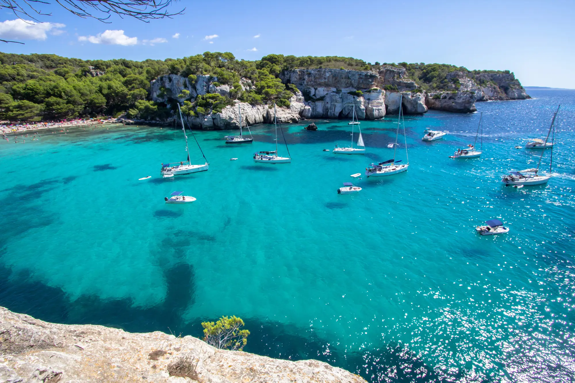 Boats and sailboats on turquoise ocean in a cala in Menorca on a sunny morning, one of the best summer destinations in Europe.