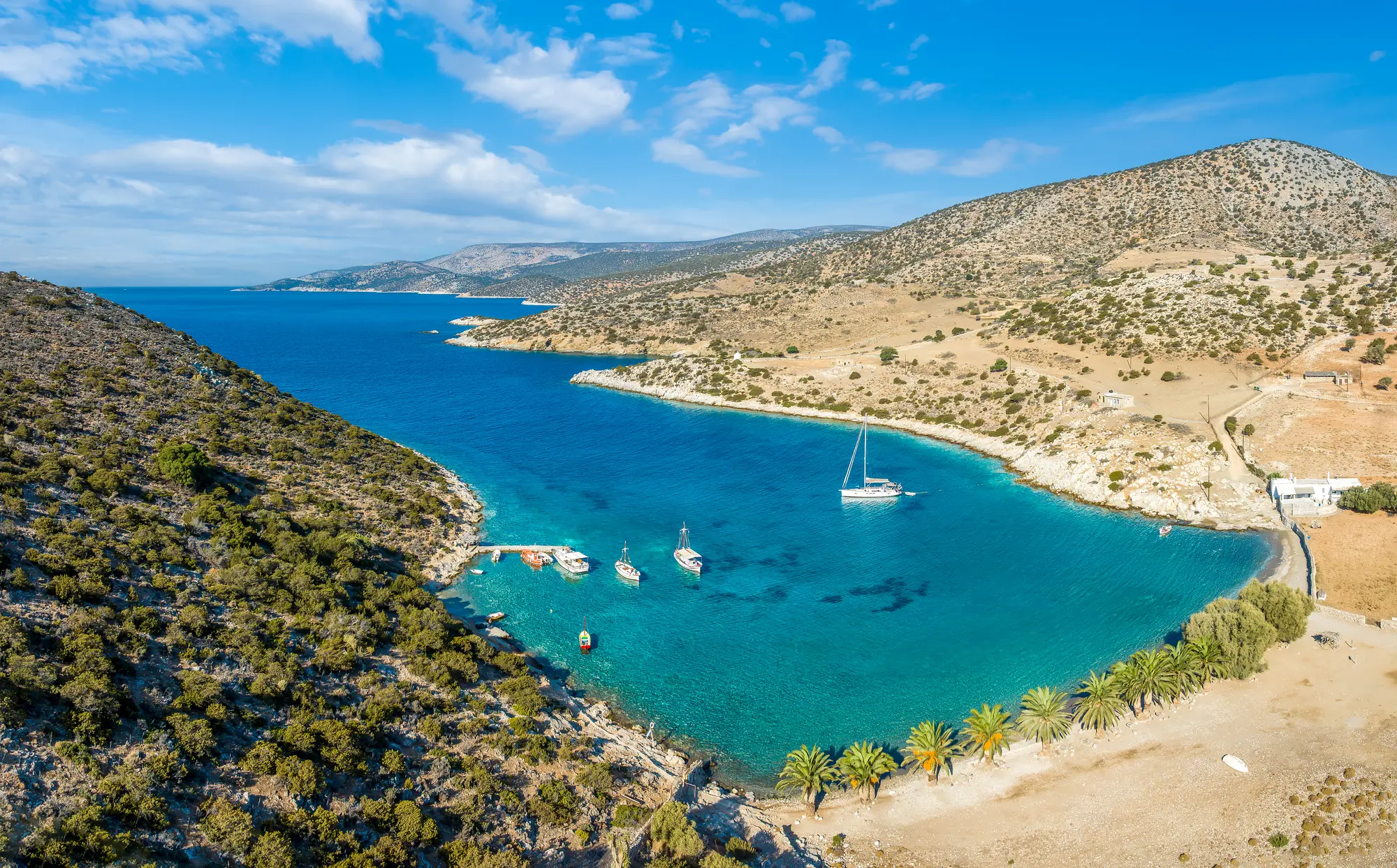 View from above of sailboats on turquoise waters by a beach with palm trees with headlands on both sides on Naxos Greece, one of the best summer destinations in Europe.