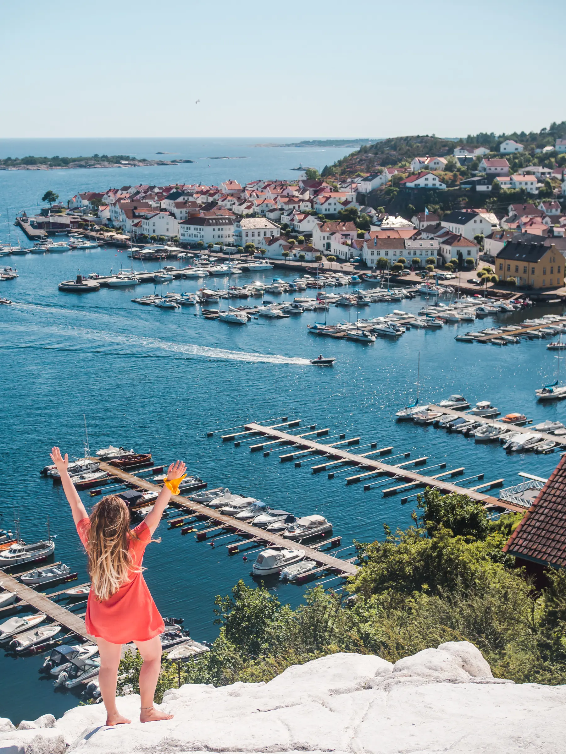 Woman with long hair, wearing a coral dress, standing with her hands in the air looking down on Risør Harbor in Norway.