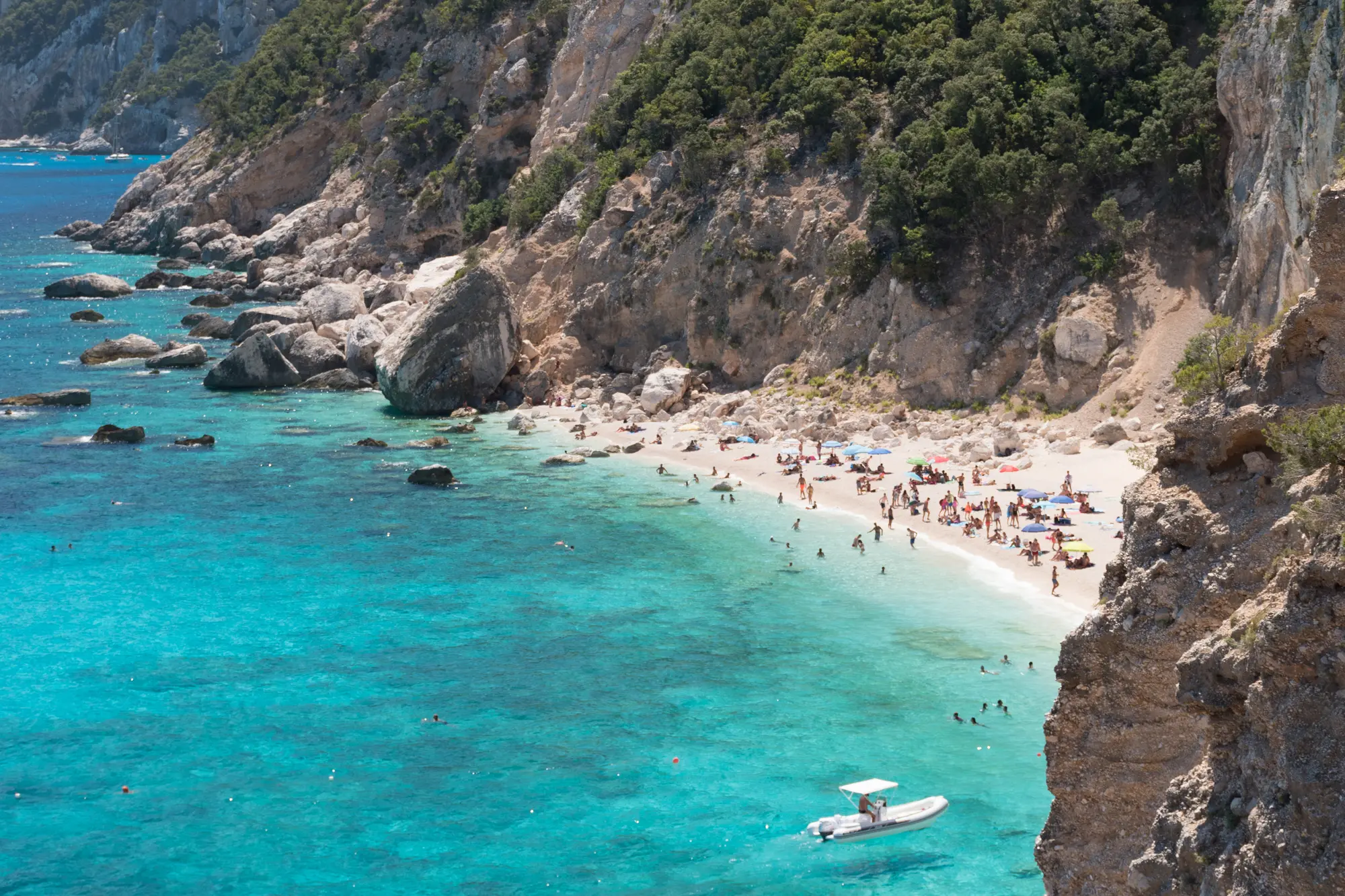 White sand beach at the bottom of a cliff with turquoise water and a white boat in Sardinia, one of the best summer destinations in Europe.