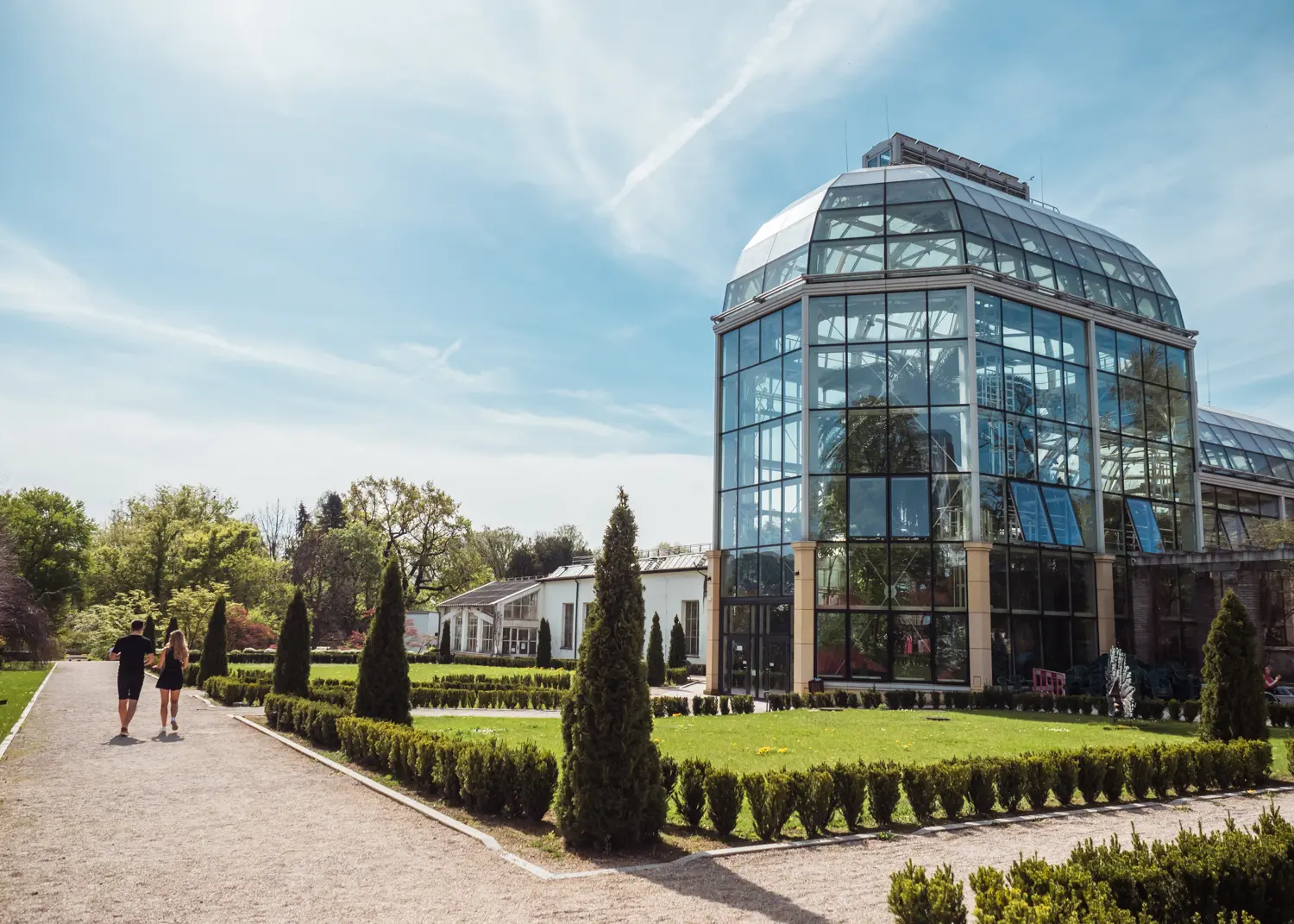 Two people walking along a gravel path surrounded by green laws and a tall greenhouse in Krakow Botanical Garden, a hidden gem in Krakow.