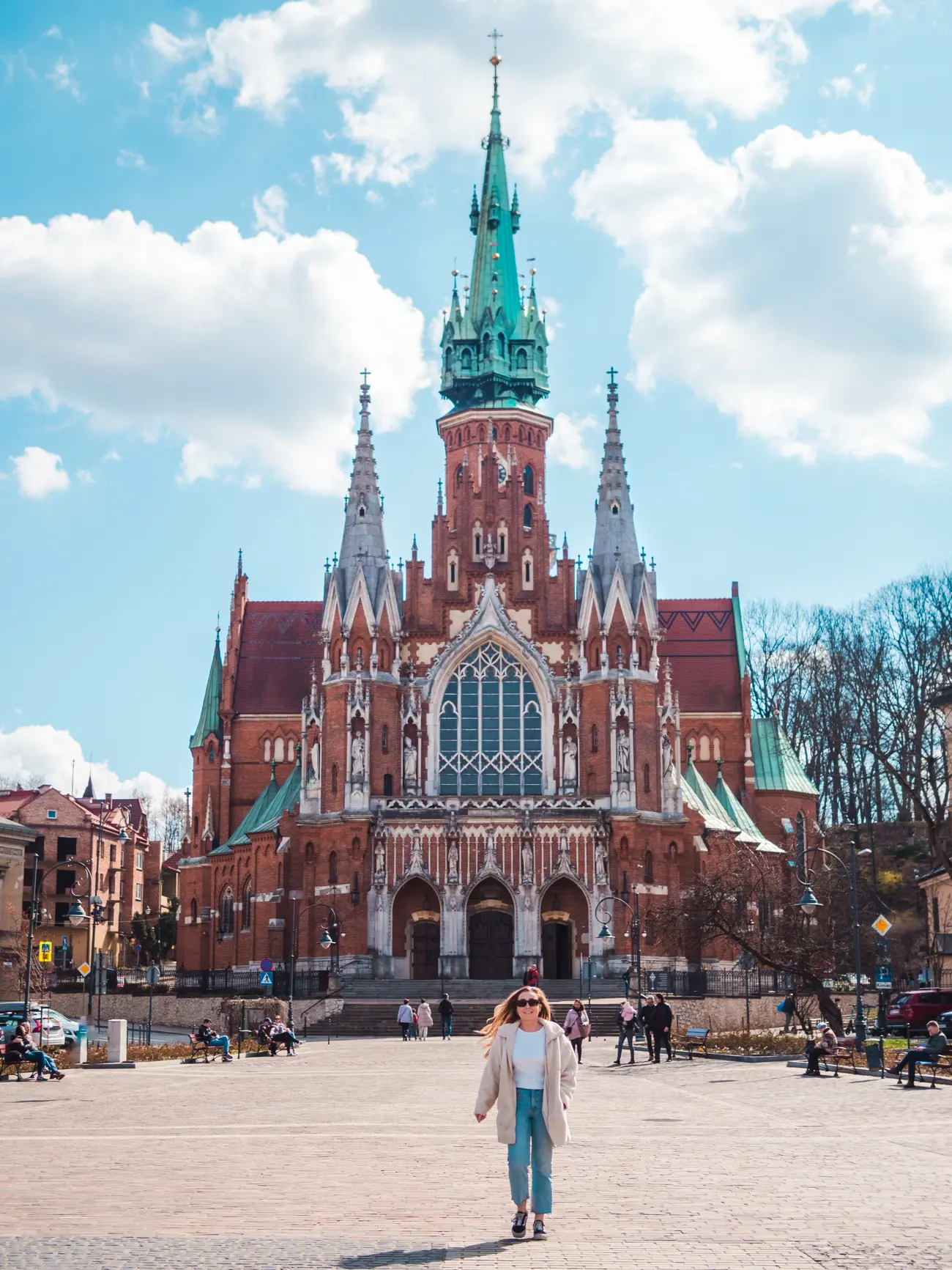 Woman wearing denim and a white coat walking across the square in front of Church of St. Joseph made from red brick with three spires, a hidden gem in Krakow.