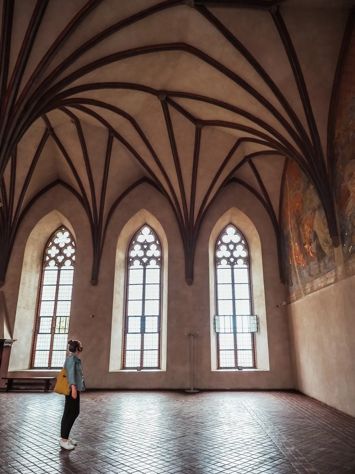 Woman wearing a denim jacket and black pants standing in the Great Refectory of Malbork Castle with the sun shining in from three tall windows.