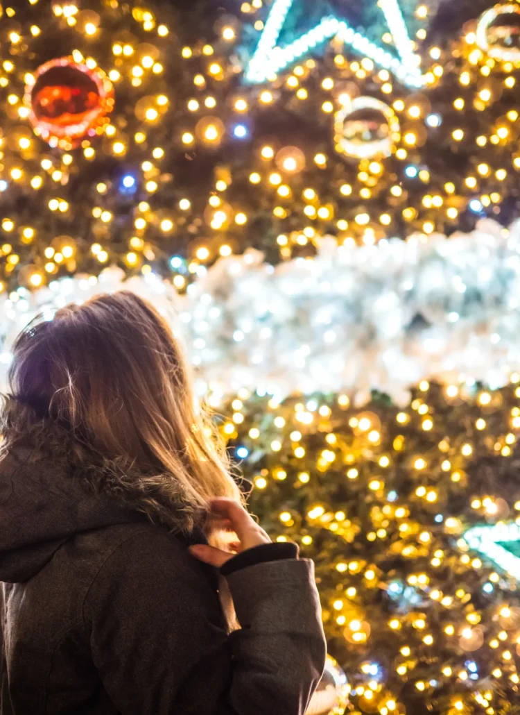 Close up of girl wearing a grey winter jacket standing in front of a huge Christmas tree decorated with thousands of light, white garland and big stars at Krakow Christmas Market.