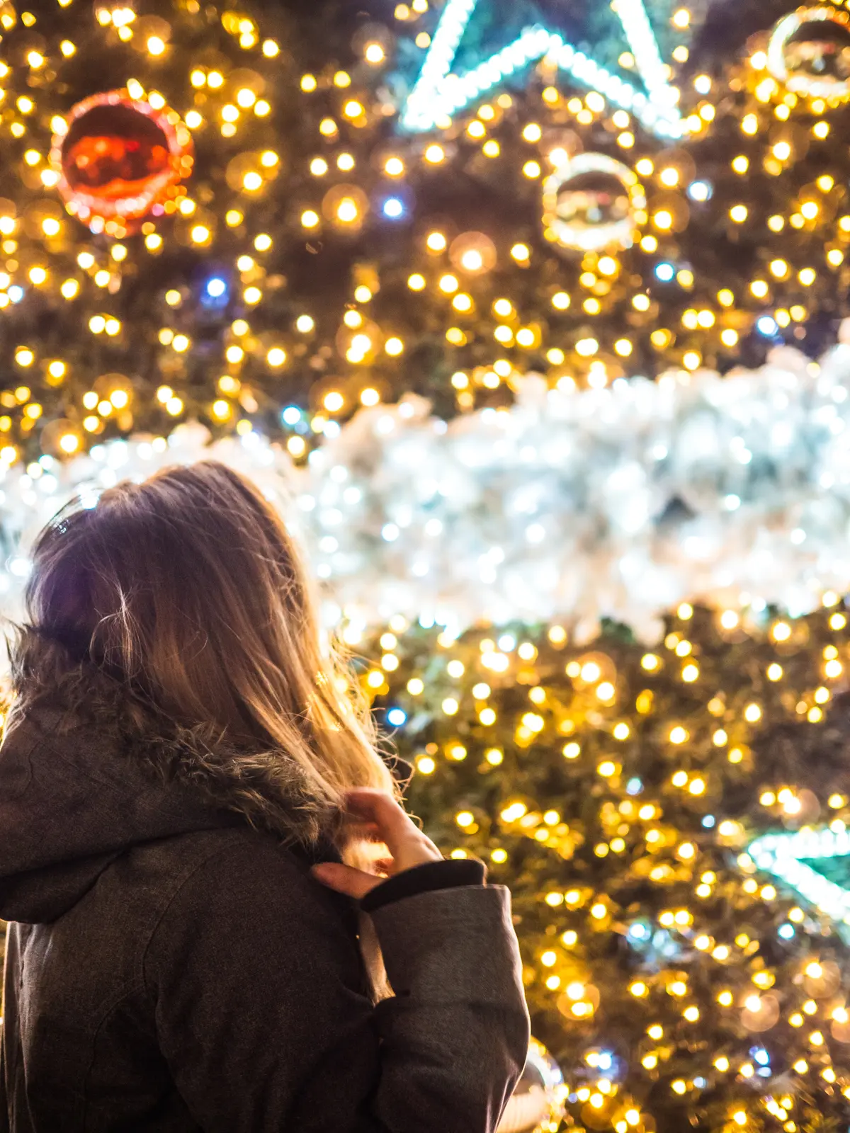 Close up of girl wearing a grey winter jacket standing in front of a huge Christmas tree decorated with thousands of light, white garland and big stars at Krakow Christmas Market.