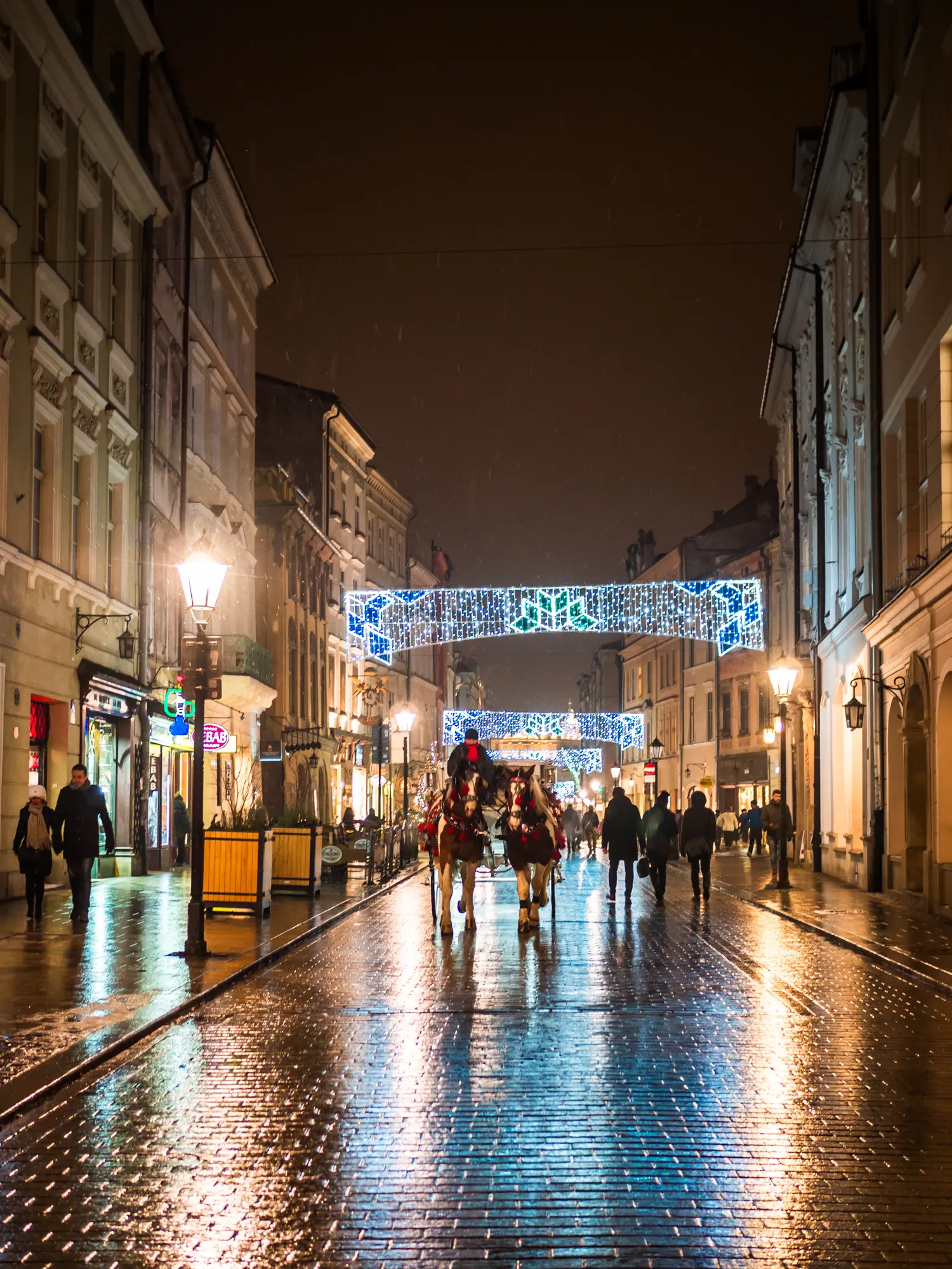 Two horses and a carriage coming down a wet cobbled street with Christmas lights in the background at Krakow Christmas Market.