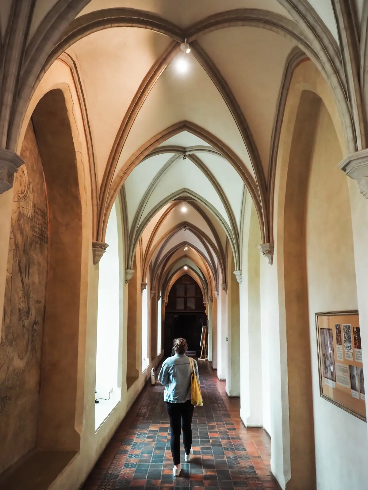 Woman wearing a denim jacket and black pants walking down a hallway with vaulted ceiling and tiles floor at Malbork Castle.