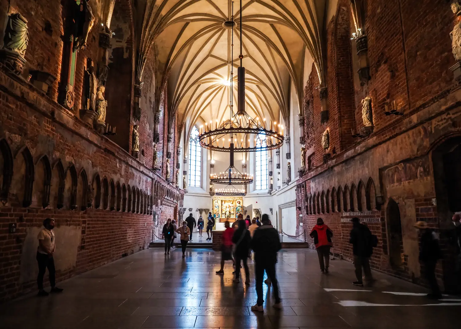 People walking under the tall vaulted ceiling with three large chandeliers in the red brick Castle Church at Malbork Castle.