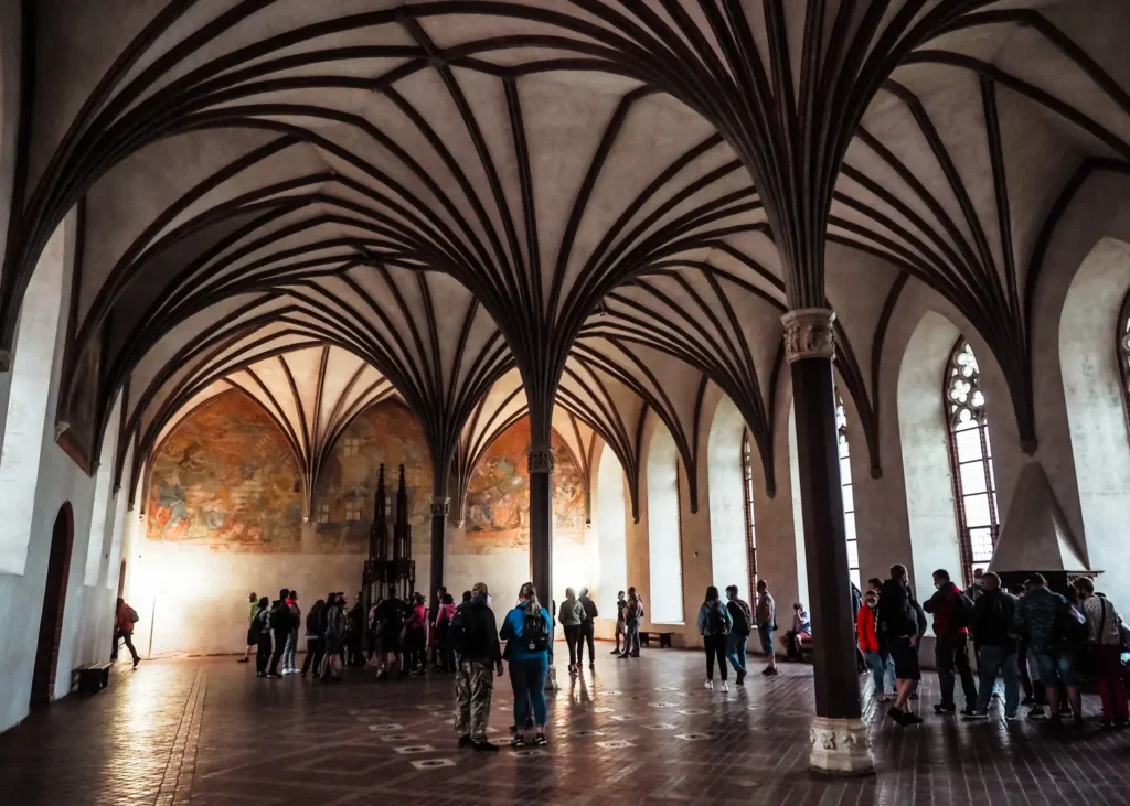 People walking in the Great Refectory of Malbork Castle with vaulted ceiling with dark brown details supported by three pillars.