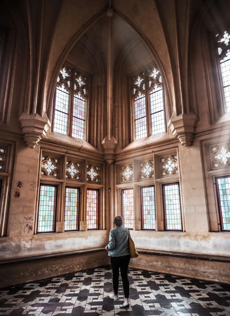 Woman wearing black pants and a denim jacket standing in the middle of a church like room with a patterned tile floor and tall ornate windows with the sun shining in at Malbork Castle.