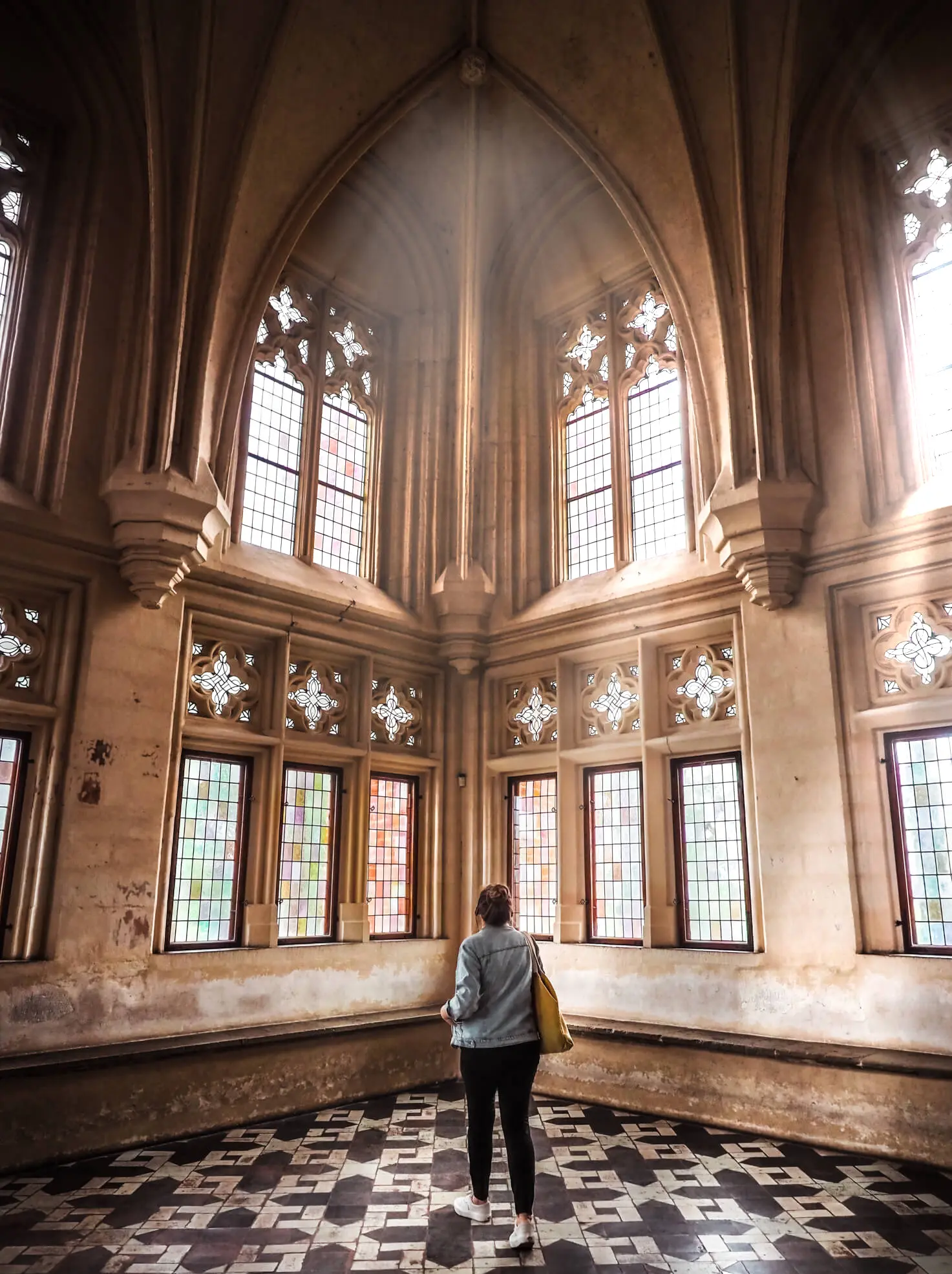 Woman wearing black pants and a denim jacket standing in the middle of a church like room with a patterned tile floor and tall ornate windows with the sun shining in the Summer Refectory at Malbork Castle.