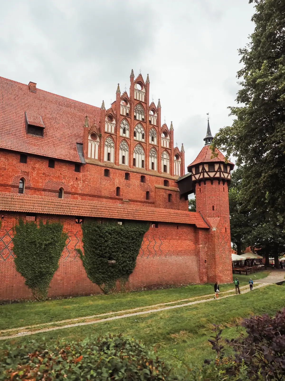 Three people walking along a path next to the massive red brick wall with tower at Malbork Castle on a day trip from Gdansk.