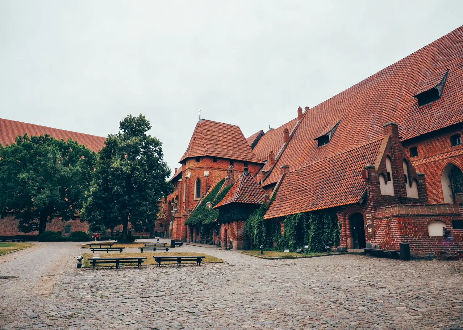 Cobbled courtyard with green trees and a seating area in the middle of the massive red brick courtyard of Malbork Castle in Poland.