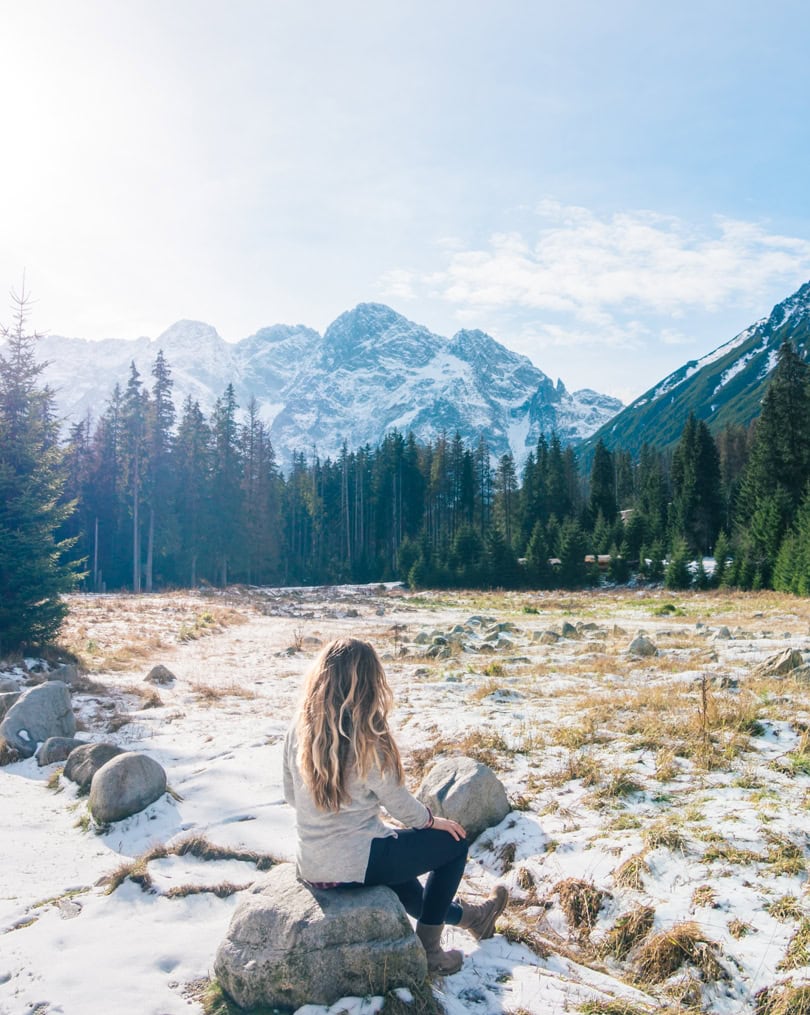 Woman with long hair wearing a grey sweater and black pants, sitting on a rock on the way to Morskie Oko Lake, mountains in the background.