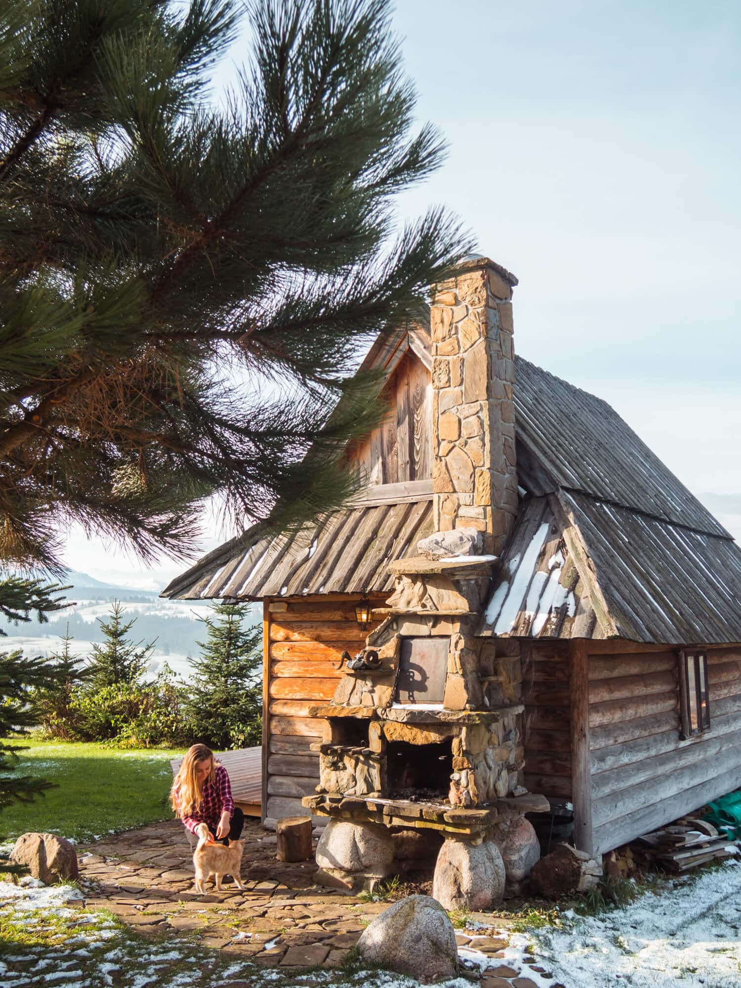 Woman with long hair wearing a purple plaid shirt sitting down next to a cat and a traditional small Polish mountain cabin, best place to stay close to Morskie Oko Lake.