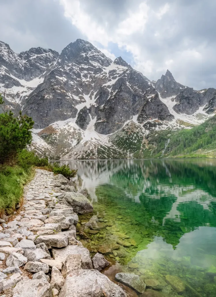 Stone path along the green Morskie Oko lake in the Tatra Mountains surrounded by mountains with some snow and greenery. Morskie Oko hiking guide.