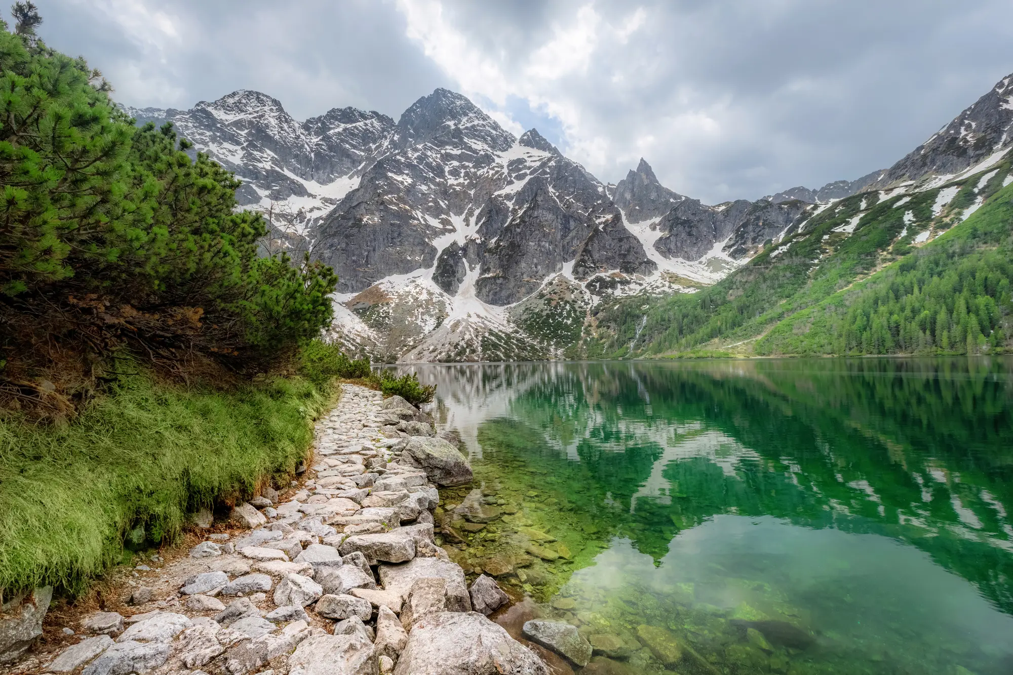 Stone path along the green Morskie Oko lake in the Tatra Mountains surrounded by mountains with some snow and greenery.