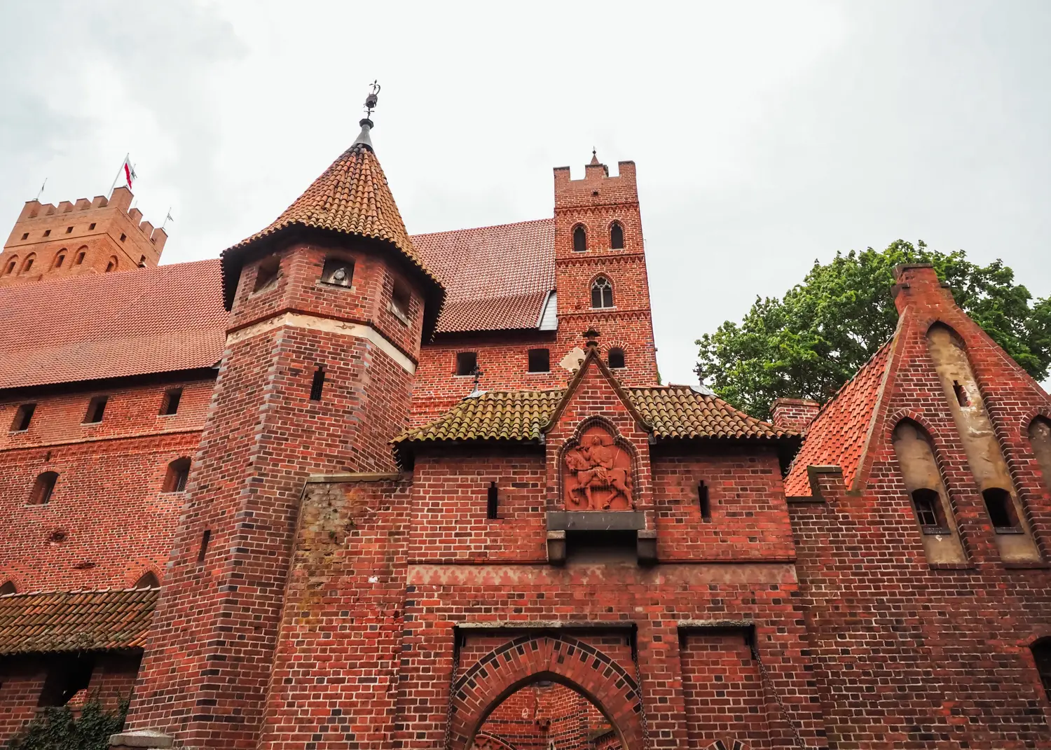 Exterior view of the red brick Malbork Castle with two square towers, one with six sides and many small windows.