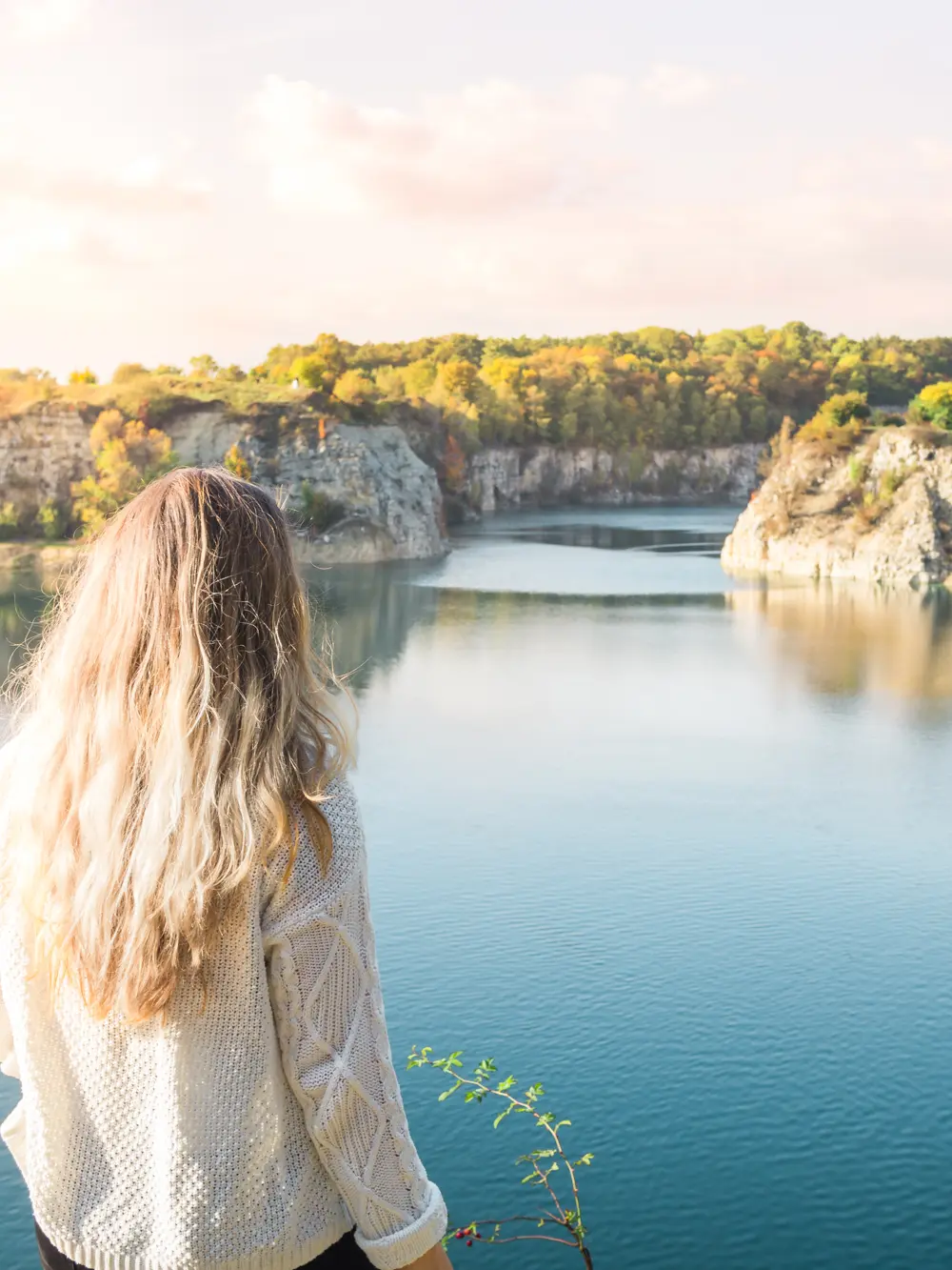 Woman with long hair wearing a white knit sweater looking out over a blue lake with cliffs covered in greenery in the background, Zakrzówek a hidden gem.