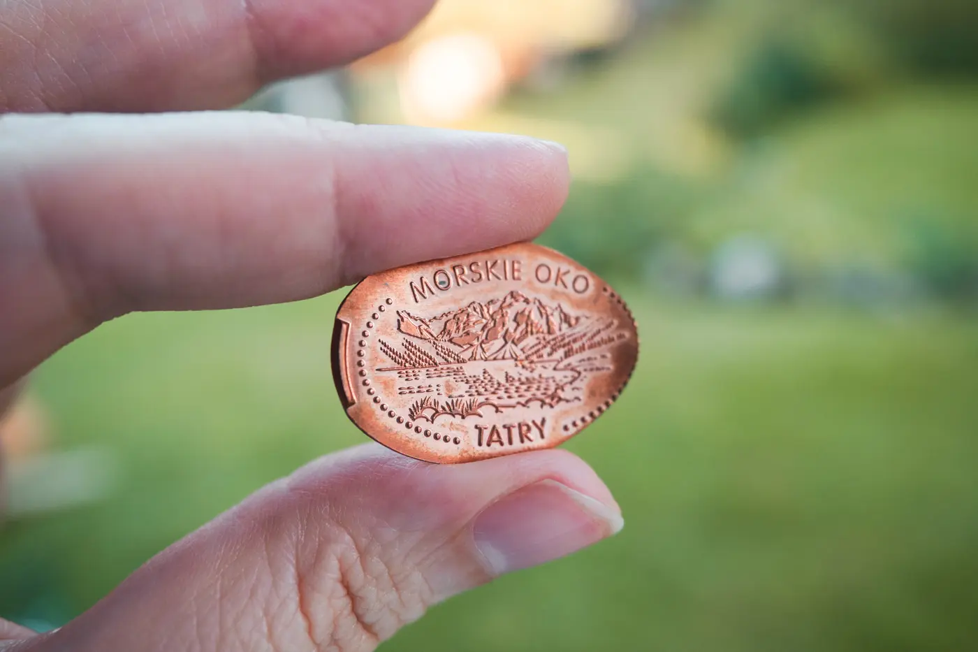 Close up of two fingers holding a copper colored souvenir penny, with green landscape in the background at Morskie Oko in southern Poland.