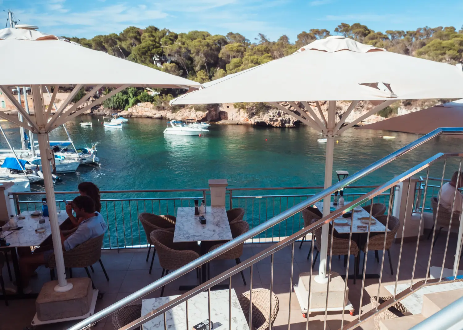View of tables, brown chairs and white parasols at Bon Bar Cala Figuera overlooking the ocean.