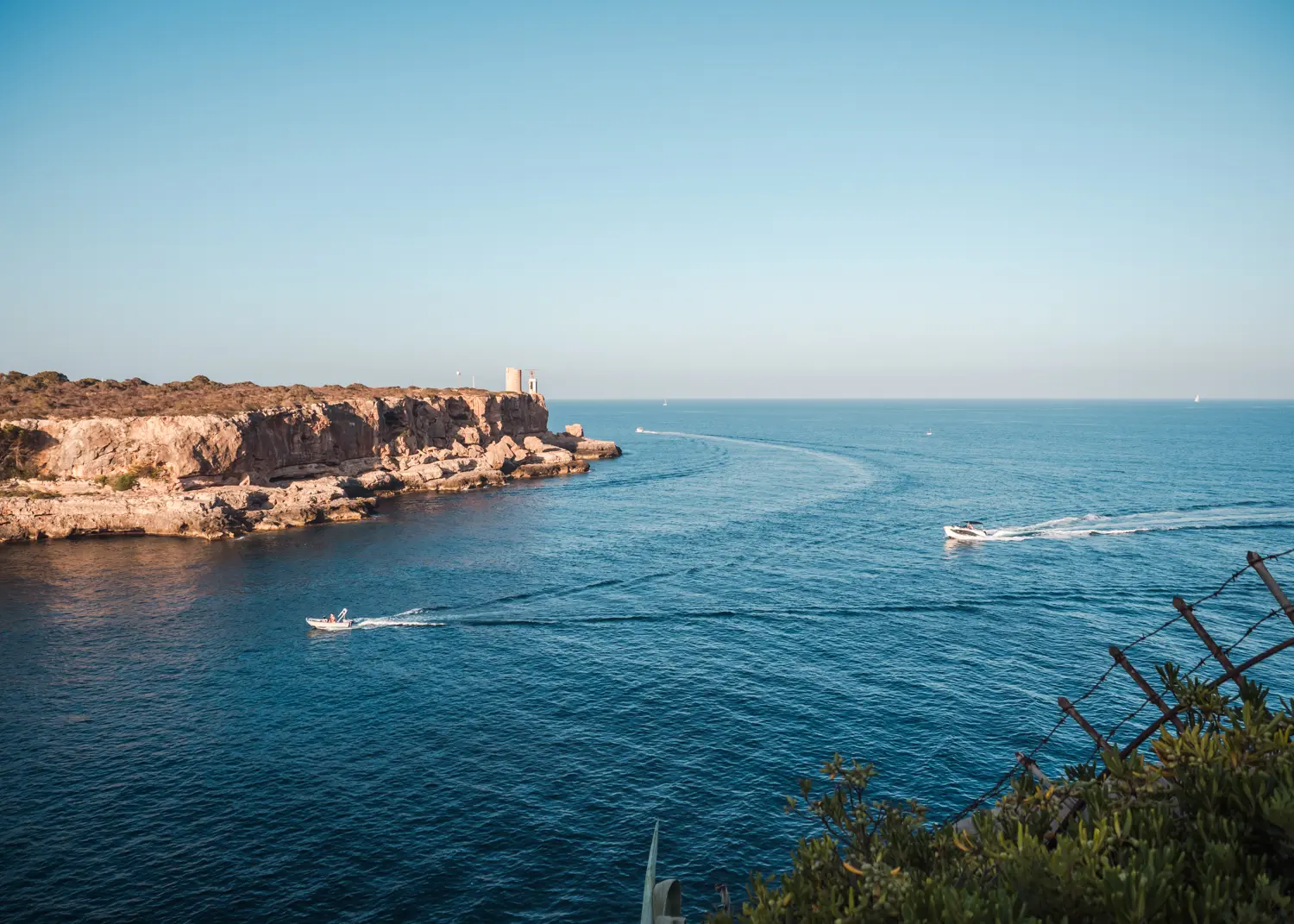 Two private boats driving past a cliff with an old small tower during sunset in Cala Figuera.