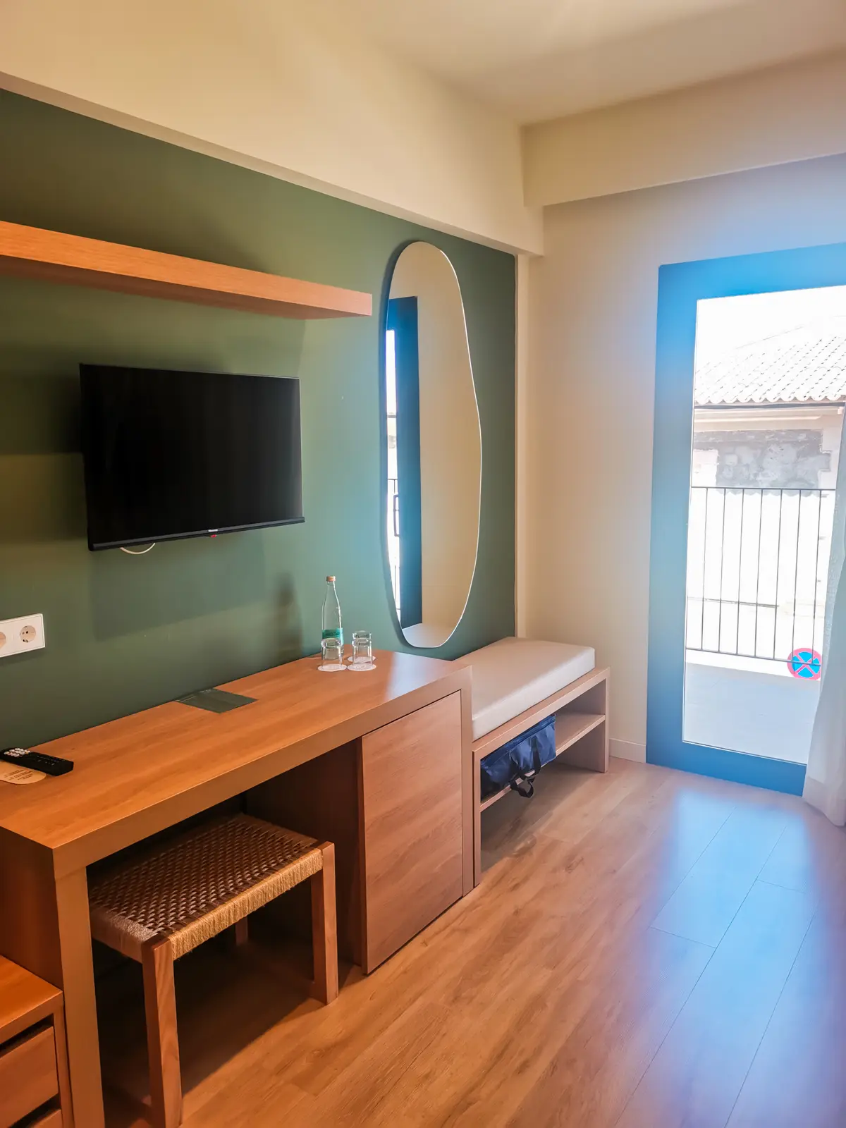 Green wall with a wooden desk and an oval mirror, sun shining in from the patio door at Hotel Santanyi Port in Cala Figuera.