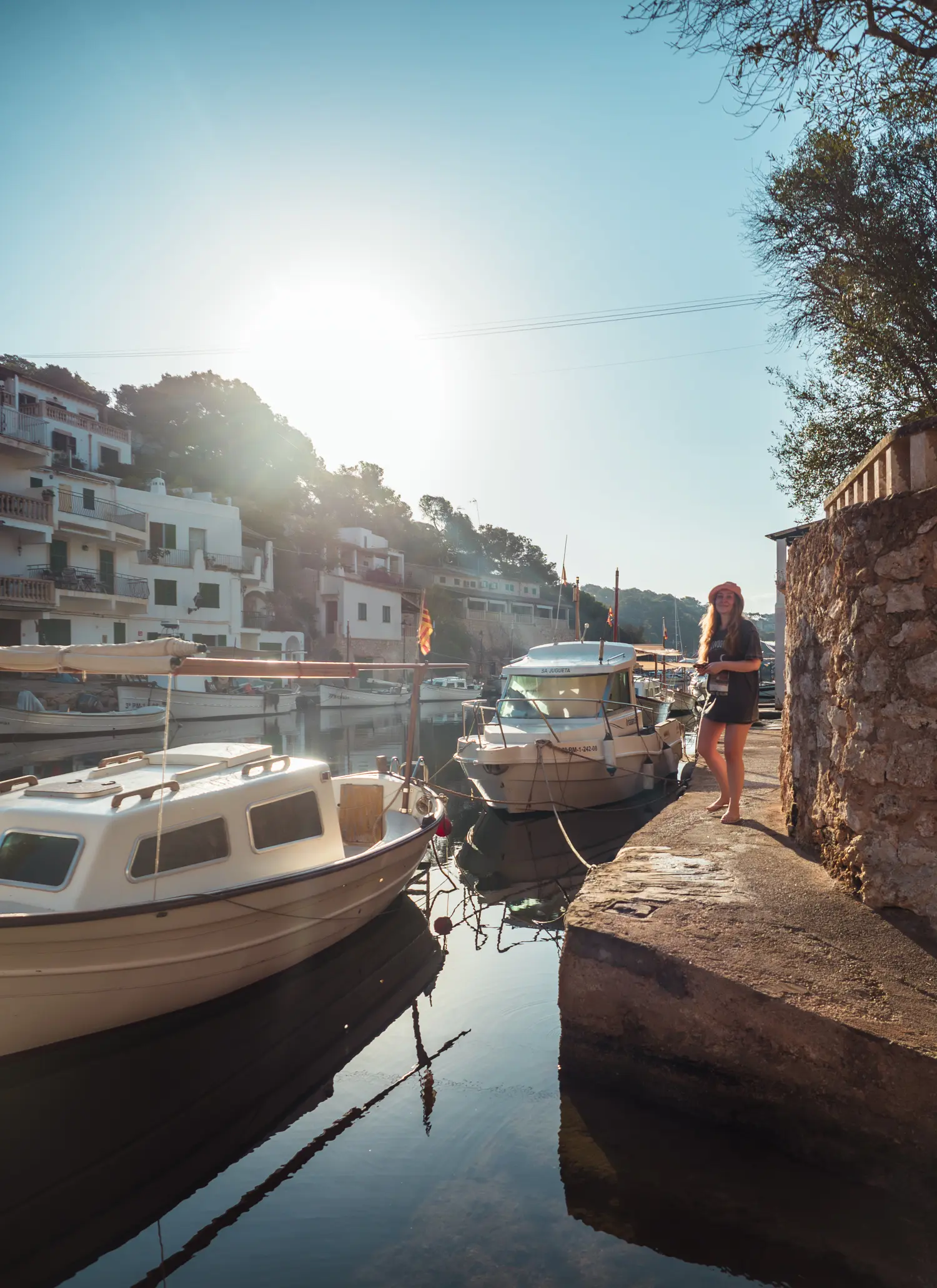 Woman with long hair, wearing a long t-shirt and bucket hat, walking along Cala Figuera Port next to two older boats with the sun shining in.