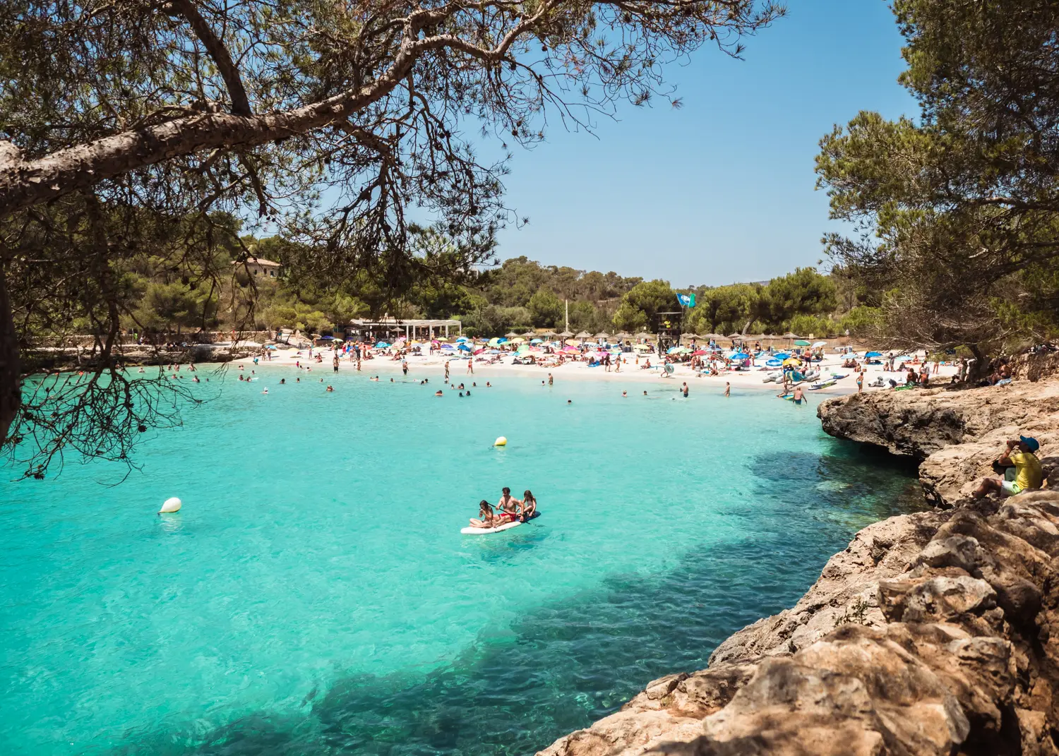 Three people on a white paddle board on the turquoise water of Cala Mondrago with a busy white beach in the background, in Mondrago Natural Park in Mallorca.