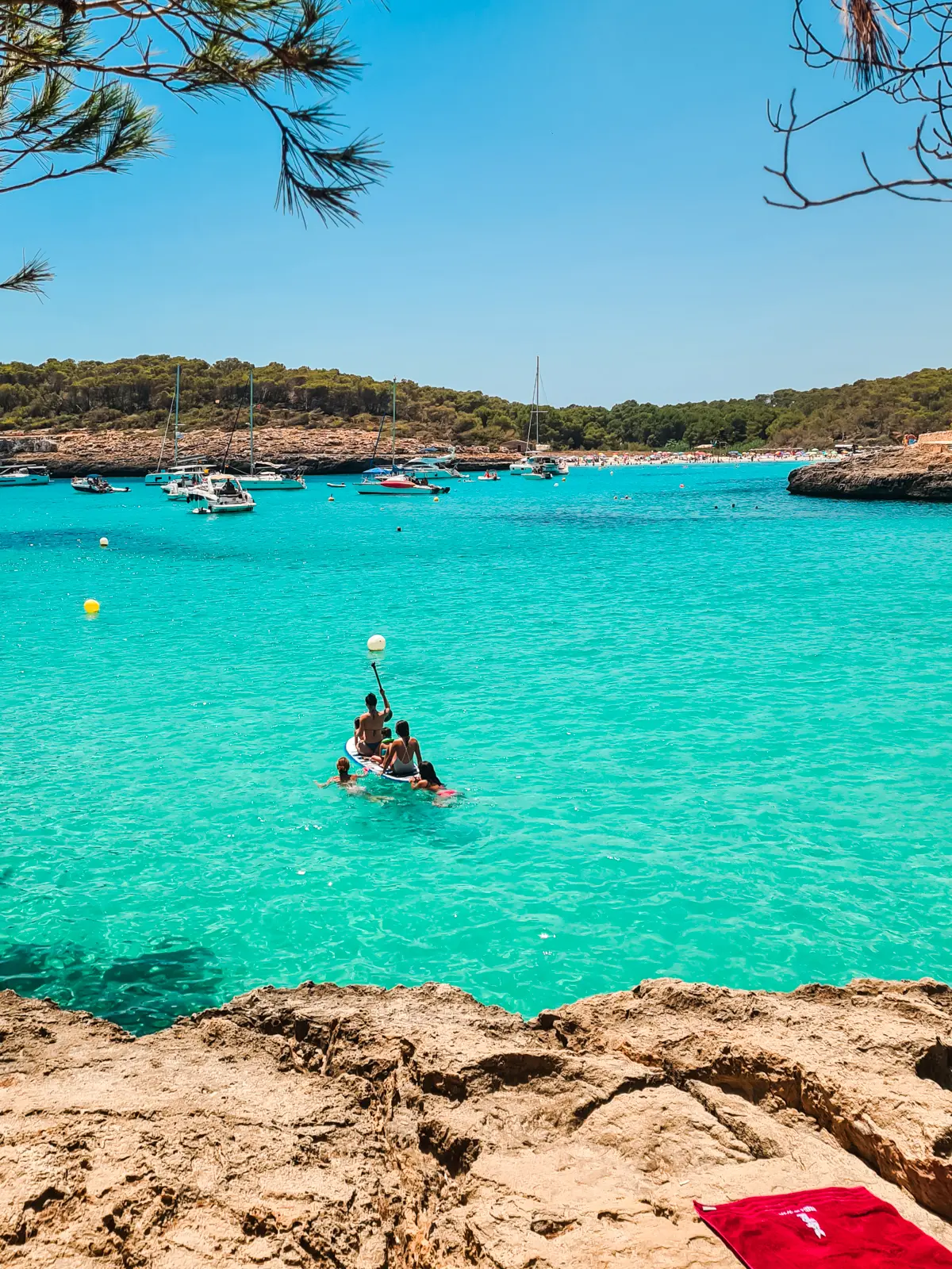 Two women and two children sitting on a paddle board in the vibrant turquoise ocean of Cala Mondrago, with sailboats and a busy Cala S'Amarador in the background.
