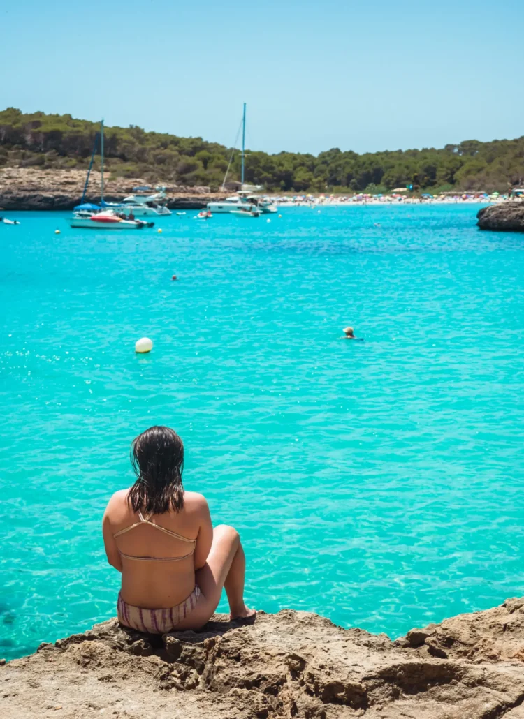 Girl with dark hair sitting on a rock looking out over the turquoise ocean of Cala Mondrago towards a busy Cala S'Amarador, two of the best beaches in Mallorca.