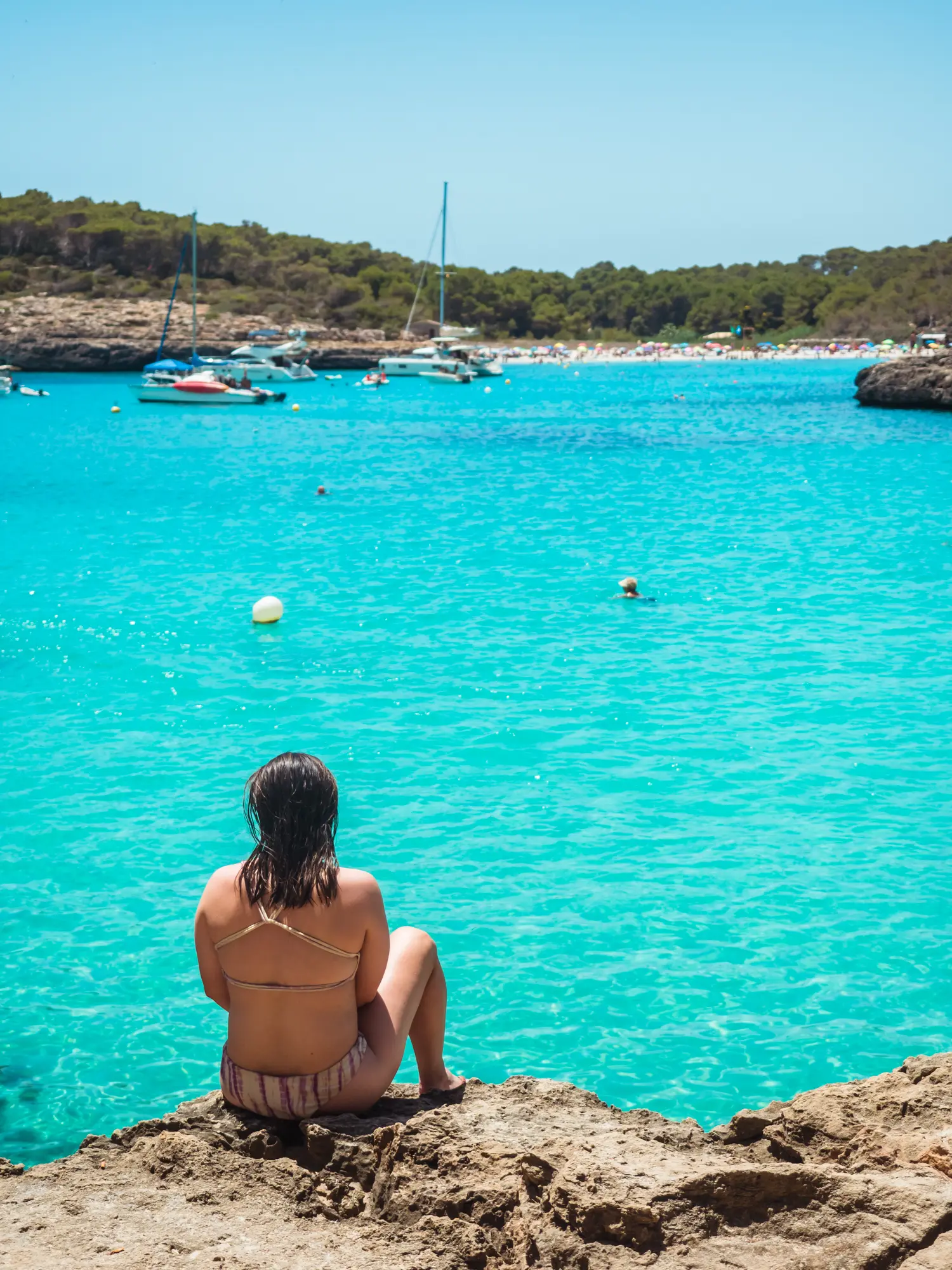 Girl with dark hair sitting on a rock looking out over the turquoise ocean of Cala Mondrago towards a busy Cala S'Amarador, two of the best beaches in Mallorca.