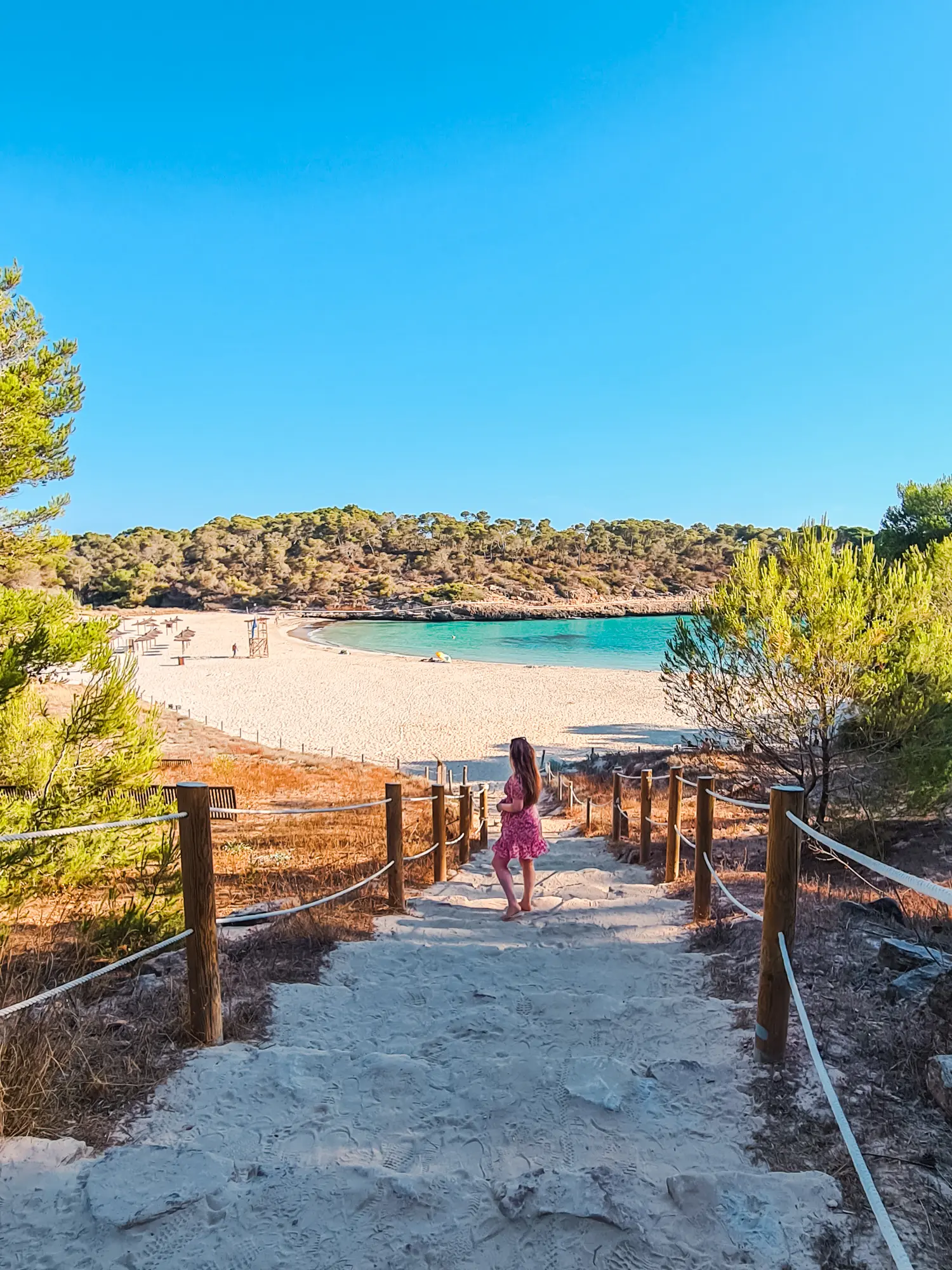Woman with long hair, wearing a pink dress, walking down sandy steps towards Cala S'Amarador surrounded by greenery and turquoise ocean.