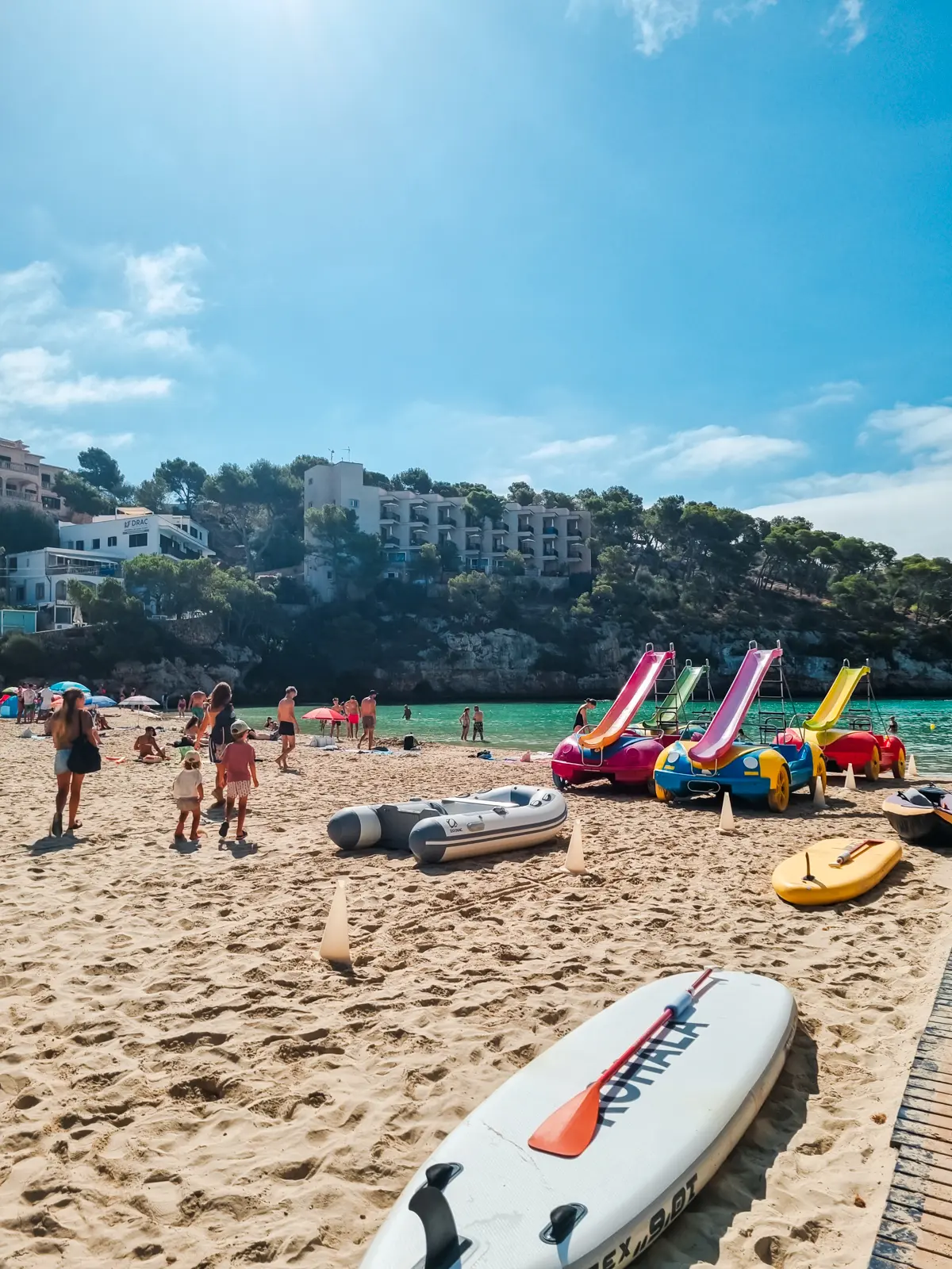 Group of people walking across Santanyi Beach with colorful boats and stand up paddle boards for rent, cliff with greenery and white hotels in the background, on a day trip from Cala Figuers.